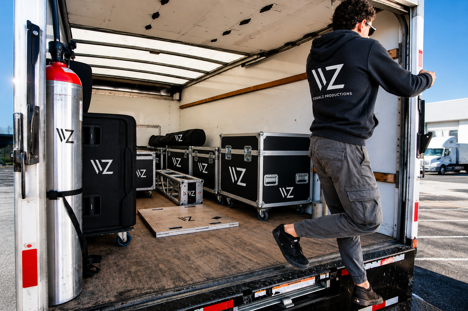 A man unloading equipment from a box truck, including cases, a fire extinguisher, and other gear branded with the 'VZ' logo.