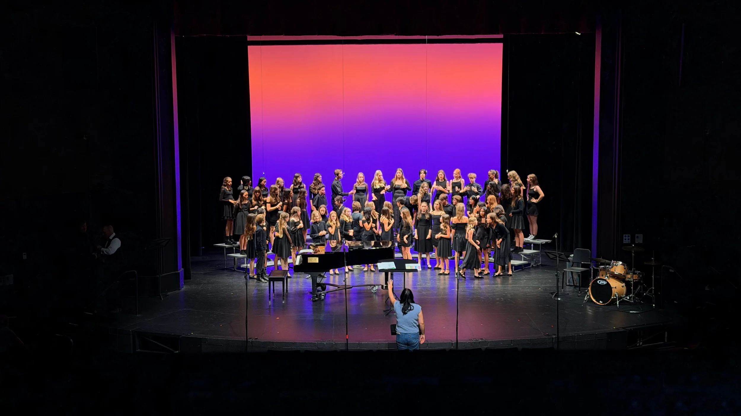 A choir of young girls dressed in black standing on risers on stage during a performance, with a colorful gradient backdrop and musical instruments including a piano, drums, and music stands nearby.