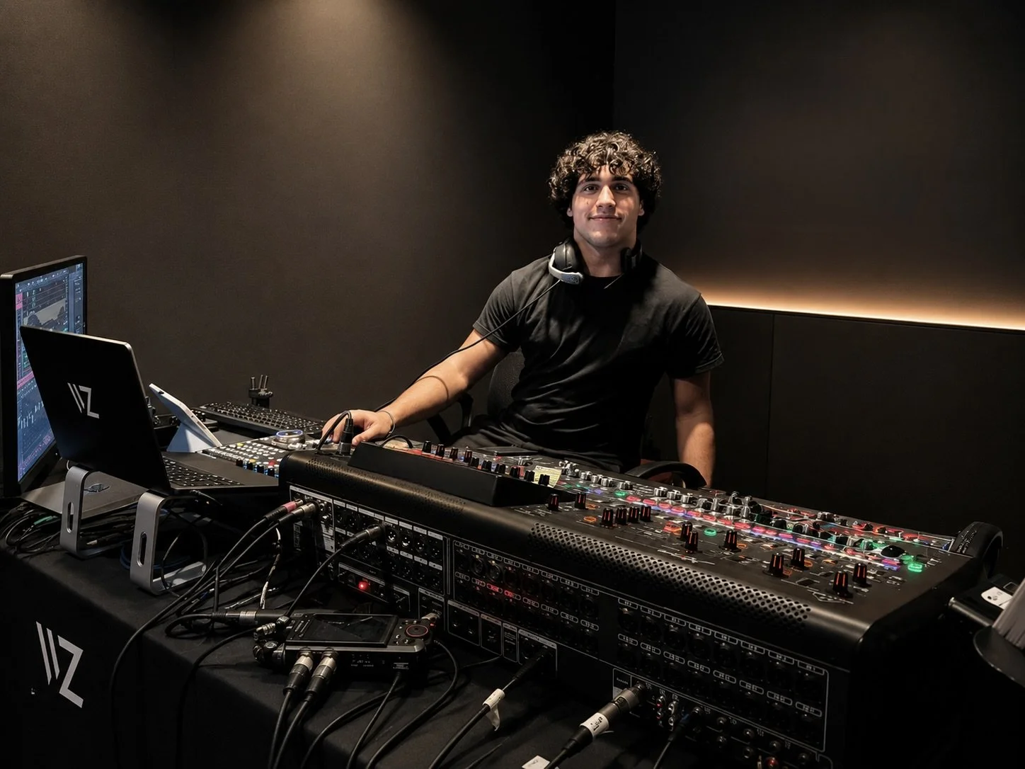 A young man with curly hair, wearing a black t-shirt, sitting at a DJ booth with sound equipment, a laptop, and headphones around his neck, inside a dimly lit room.