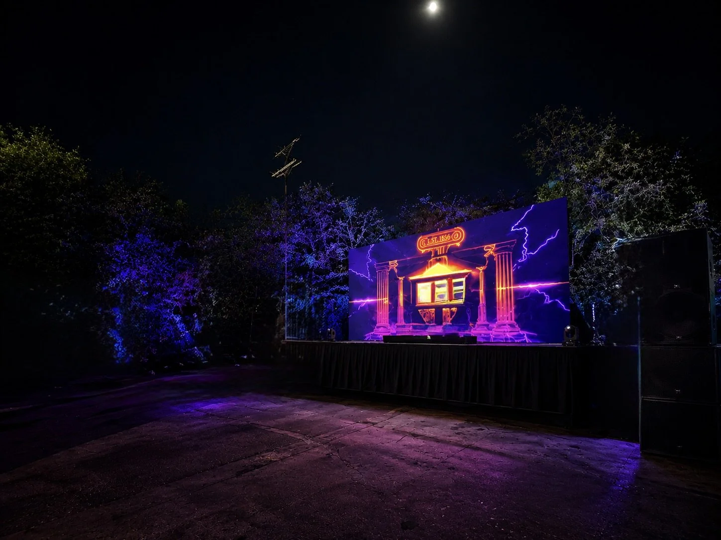 An outdoor stage with a large digital screen displaying a neon outline of a building with columns and lightning, surrounded by trees illuminated with purple and blue lights, under a night sky with the moon.