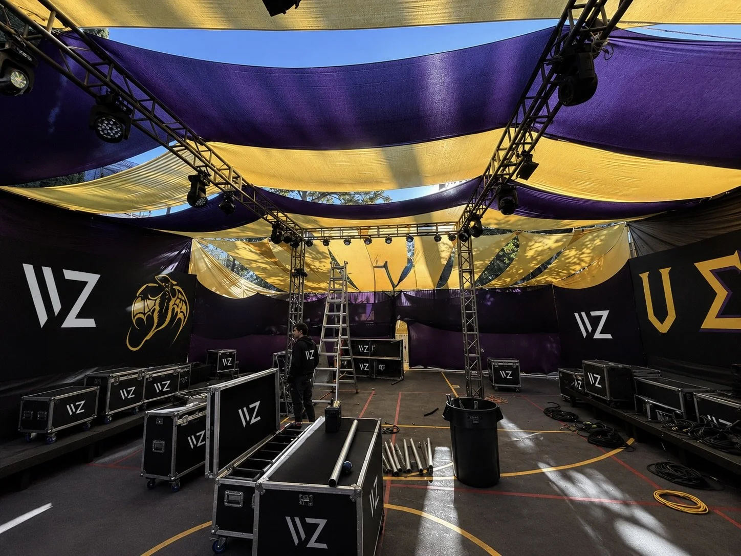 A stage setup in an outdoor area with yellow and purple fabric draped overhead. There are lighting rigs, a ladder, and black equipment cases labeled "WIZ." Black banners with a golden dragon logo and Greek letters are on each side of the stage. The scene is being prepared for an event.