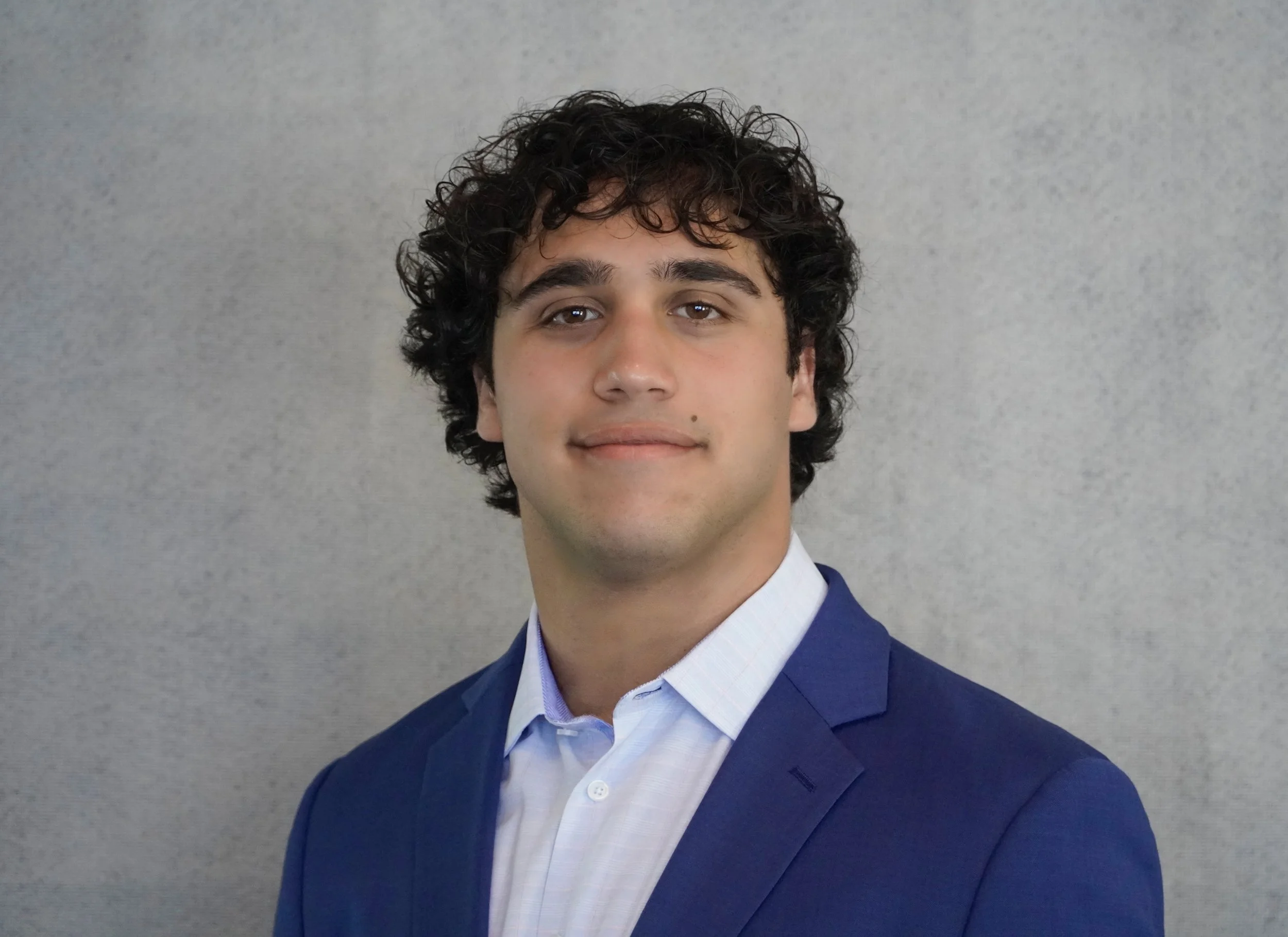 Portrait of a young man with dark curly hair, wearing a blue suit and light blue dress shirt, standing against a textured light-colored background.