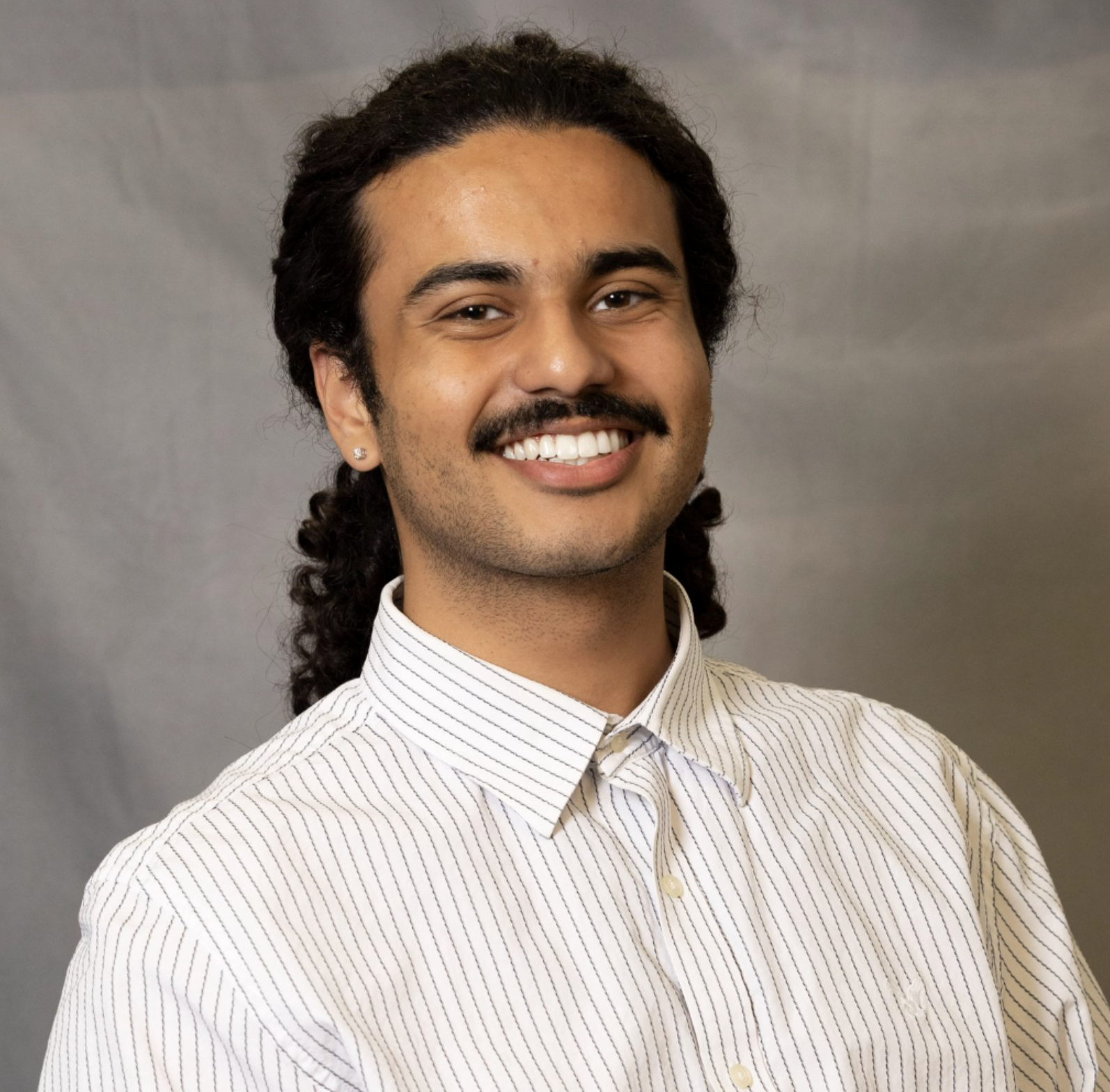 Portrait of a man with long curly black hair, a mustache, and a small beard, smiling, wearing a light-colored striped shirt, standing against a gray background.