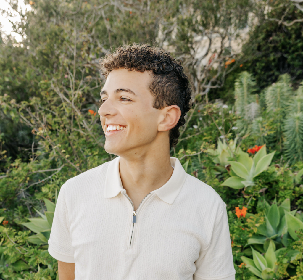 Young man with curly hair smiling outdoors among green plants and colorful flowers