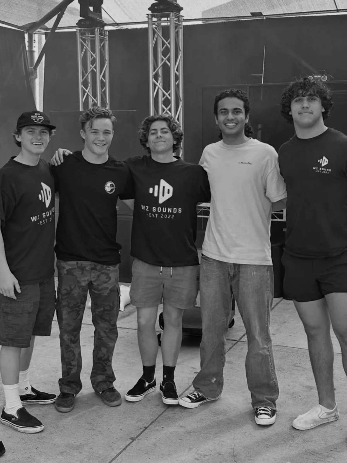 Five young men standing together indoors, smiling, with their arms around each other, in front of a stage setup with metal trusses and speakers. Some are wearing matching T-shirts with the text 'WZ SOUNDS EST 2022'.