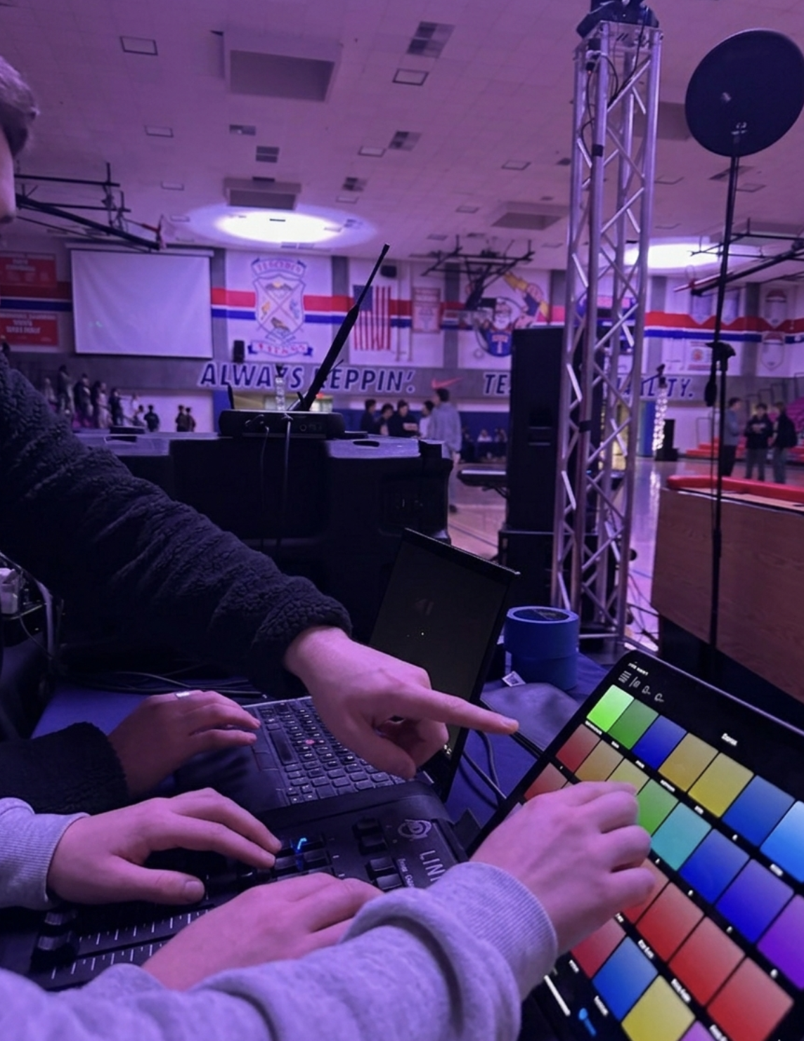 People working with equipment at a DJ or production setup inside a gymnasium or sports hall, with banners and banners with sports logos hanging on the walls.