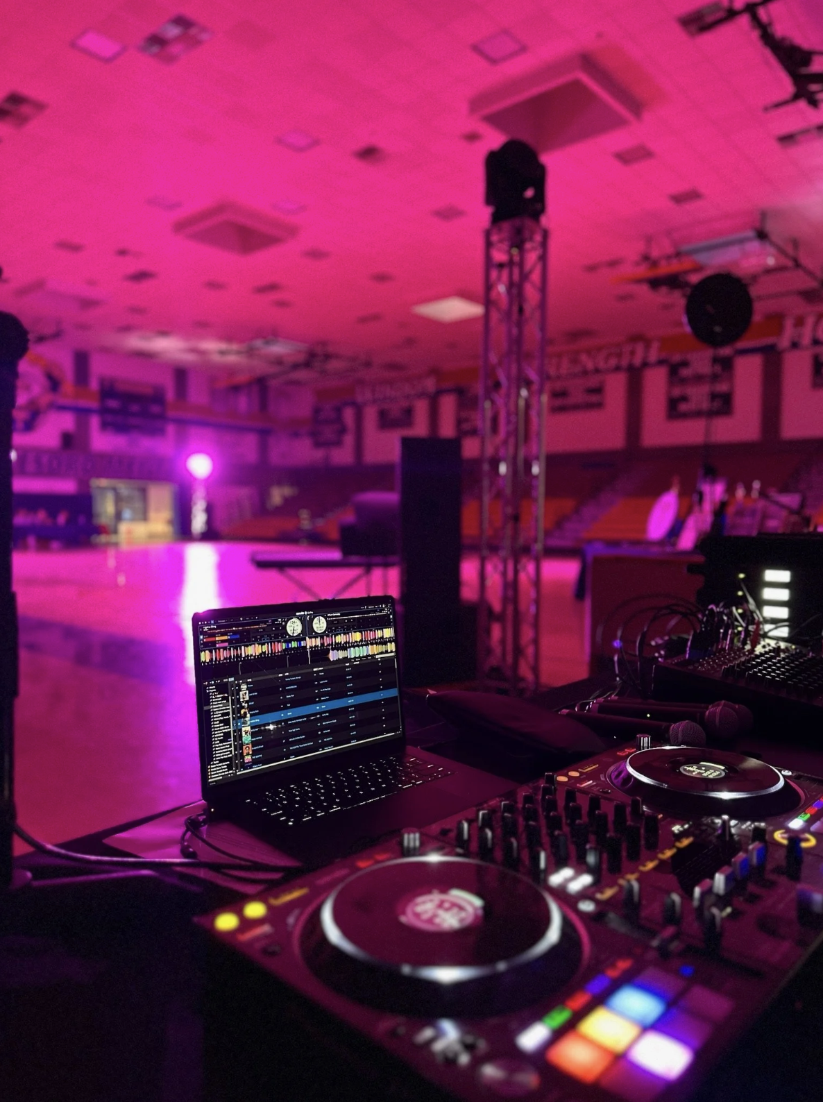DJ setup with laptop, turntables, and audio equipment in a dimly lit indoor venue with pink lighting and an empty dance floor.