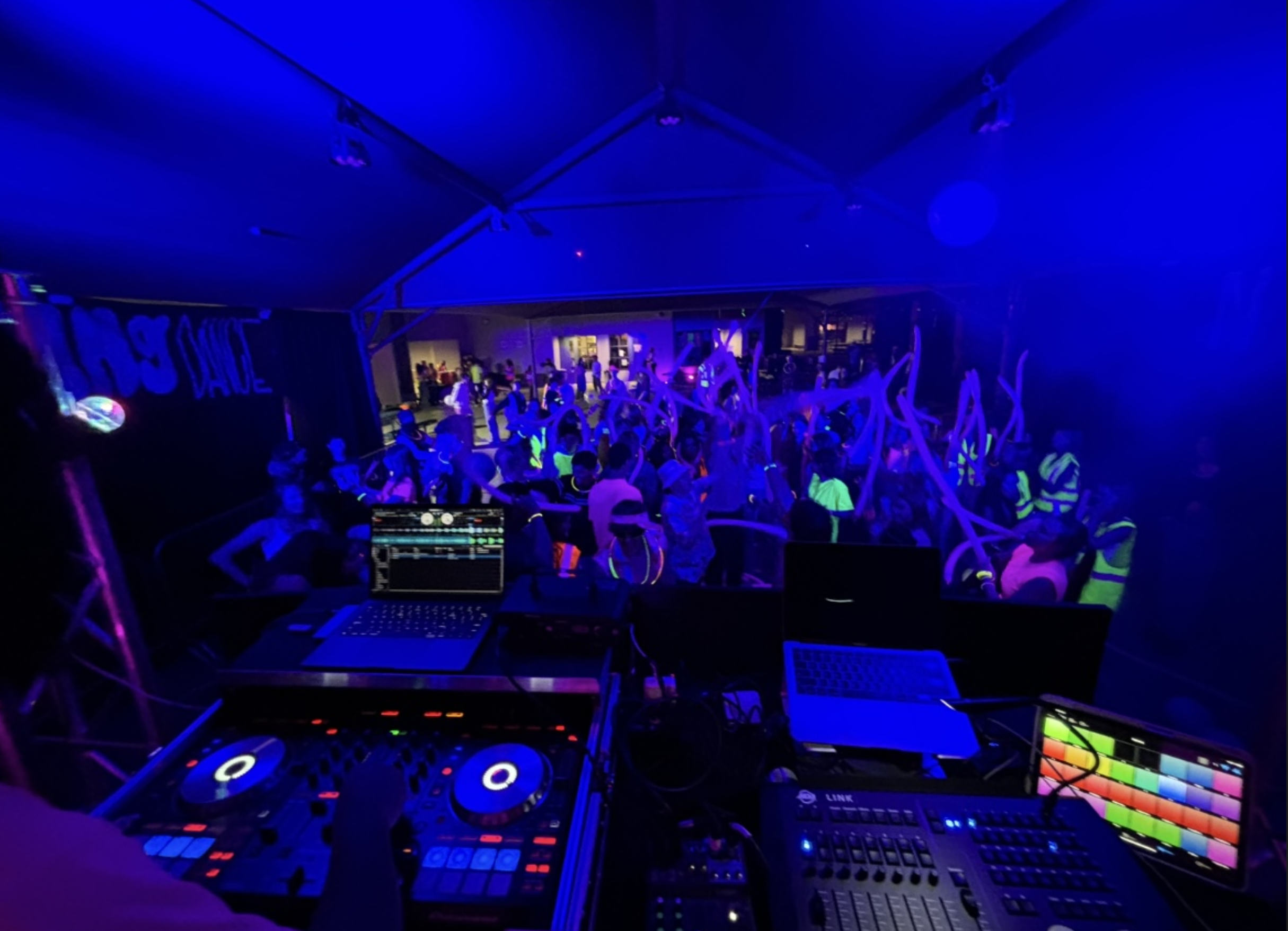 A DJ setup at a party with turntables, a mixer, and a laptop, all illuminated in purple and blue lighting, with a crowd of party-goers dancing in the background.