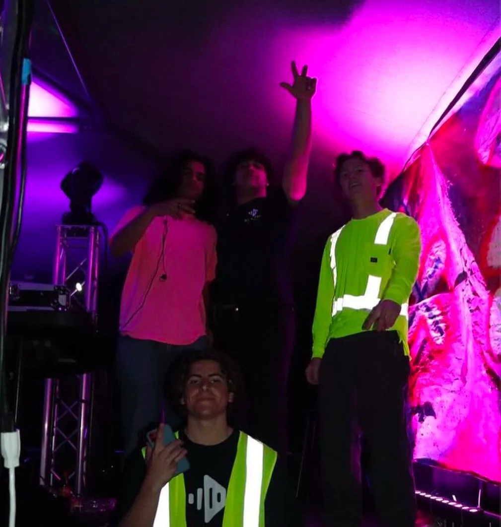 Four people on a stage with colorful purple and pink lighting, three standing and one sitting, two making peace signs, in a dark environment, possibly at a concert or event.