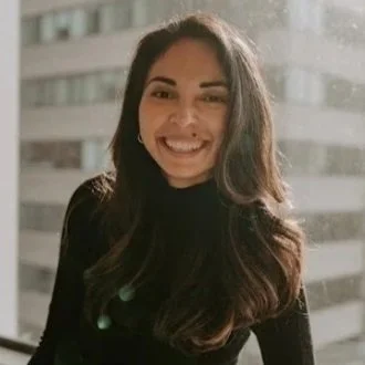A smiling woman with long wavy brown hair wearing a patterned jacket and a green top, standing in front of green tropical plants.