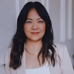 A woman with short brown hair laughing and clapping, wearing a black leather jacket, black top, gray skirt, and layered necklaces, standing against a plain gray background.