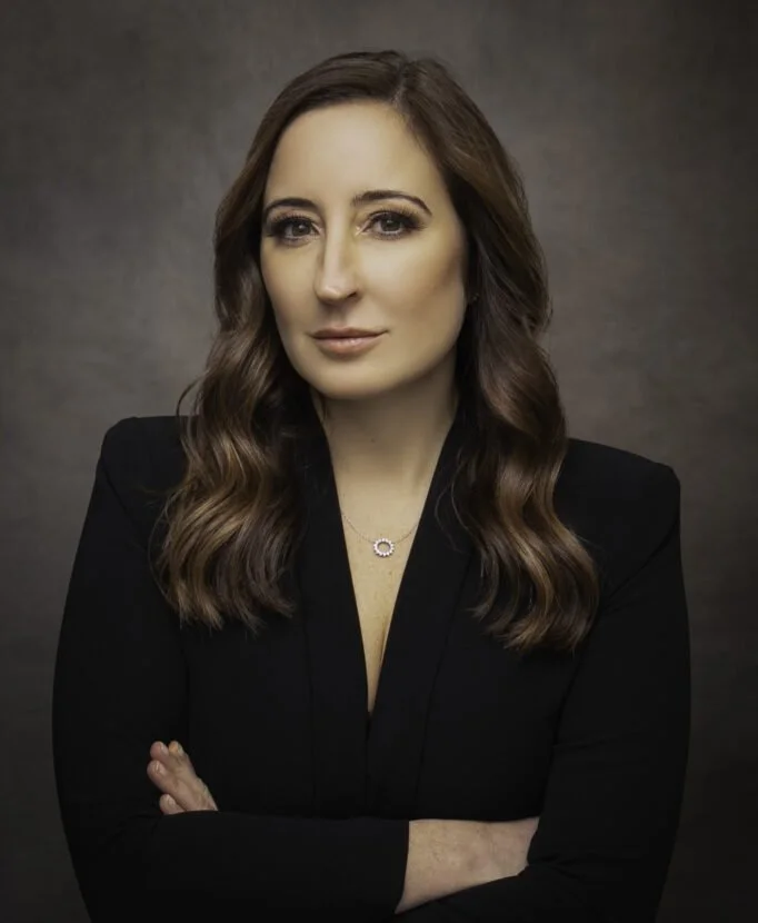 Black and white portrait of a woman with long dark hair, smiling, wearing a leather jacket and a white shirt, sitting with one leg crossed, against a plain background.
