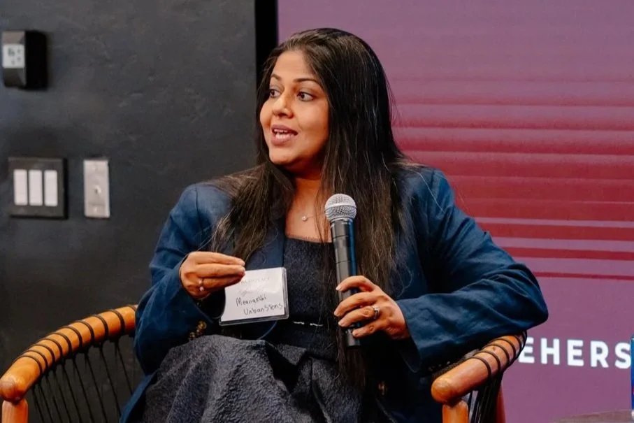A young woman with long dark hair, wearing a black top and layered necklaces, looking at the camera with a neutral expression, against a plain white background.