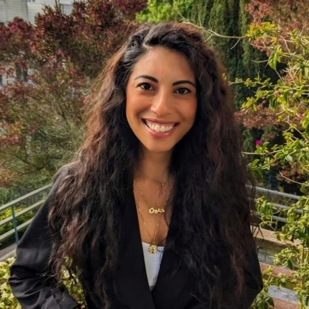 A woman with black wavy hair and a white sleeveless top leaning against a wall outdoors, smiling at the camera.