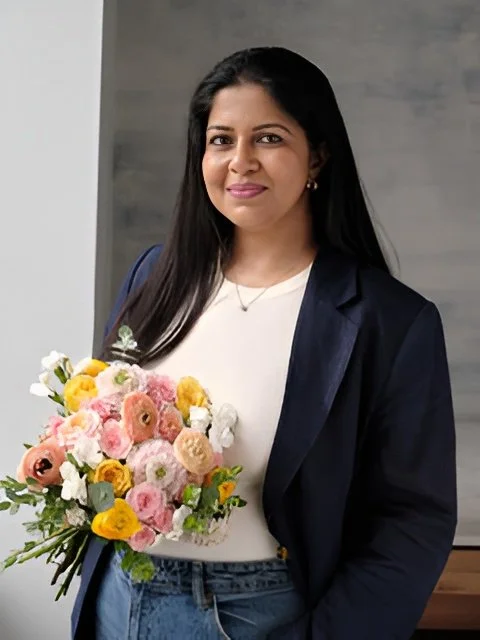 A young woman with long dark hair, wearing a black top and layered necklaces, looking at the camera with a neutral expression, against a plain white background.