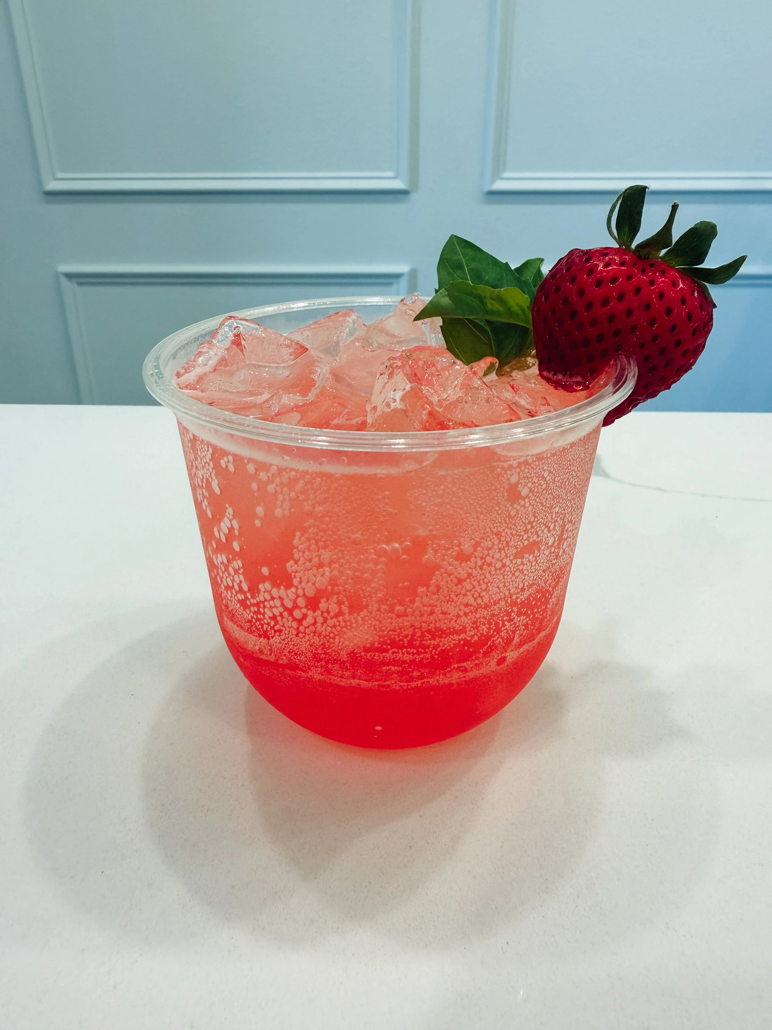 A clear plastic cup filled with pink lemonade and ice, garnished with two strawberries and a green leaf, on a white surface with a blue wall in the background.