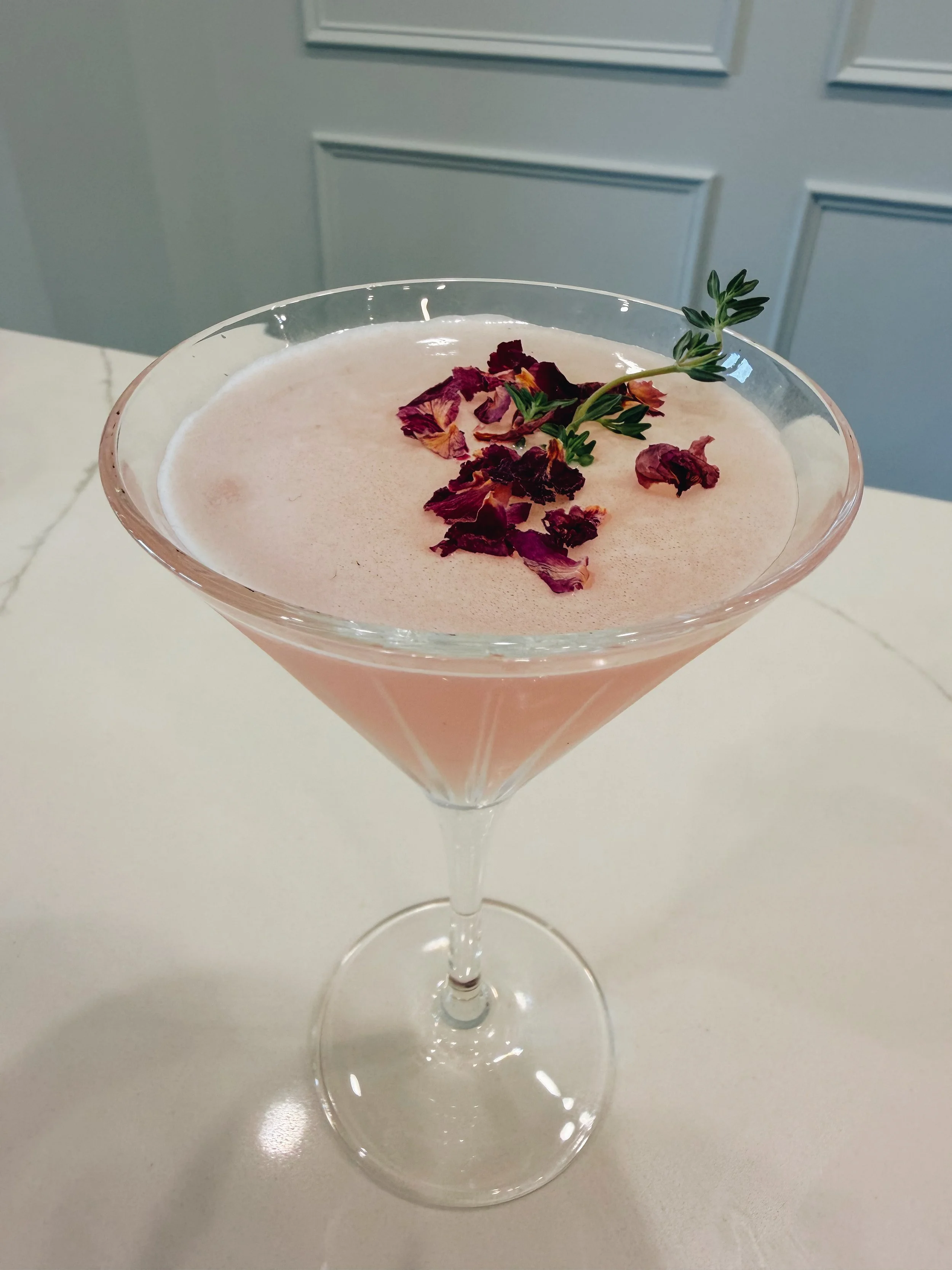 A pink cocktail garnished with dried rose petals and a sprig of thyme in a martini glass.