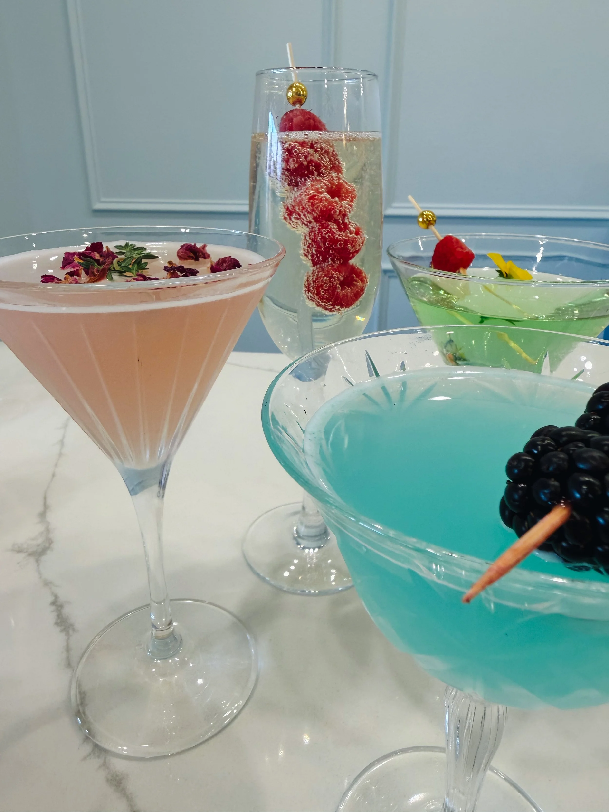 Assorted colorful cocktails on a white marble surface with a gray wall background.