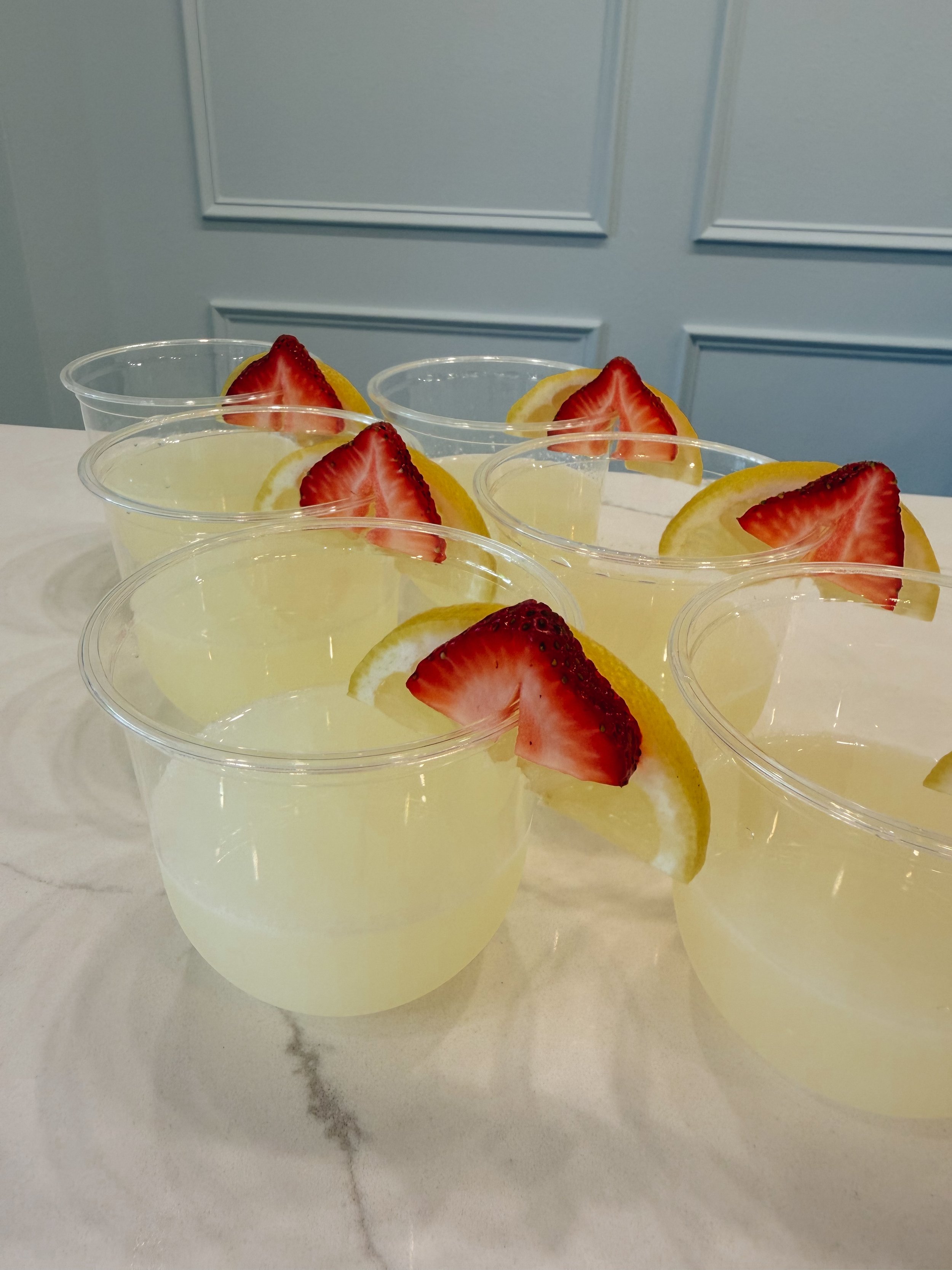 Six plastic cups filled with lemonade, each garnished with a wedge of lemon and a slice of strawberry, on a white marble surface with a light blue wall in the background.
