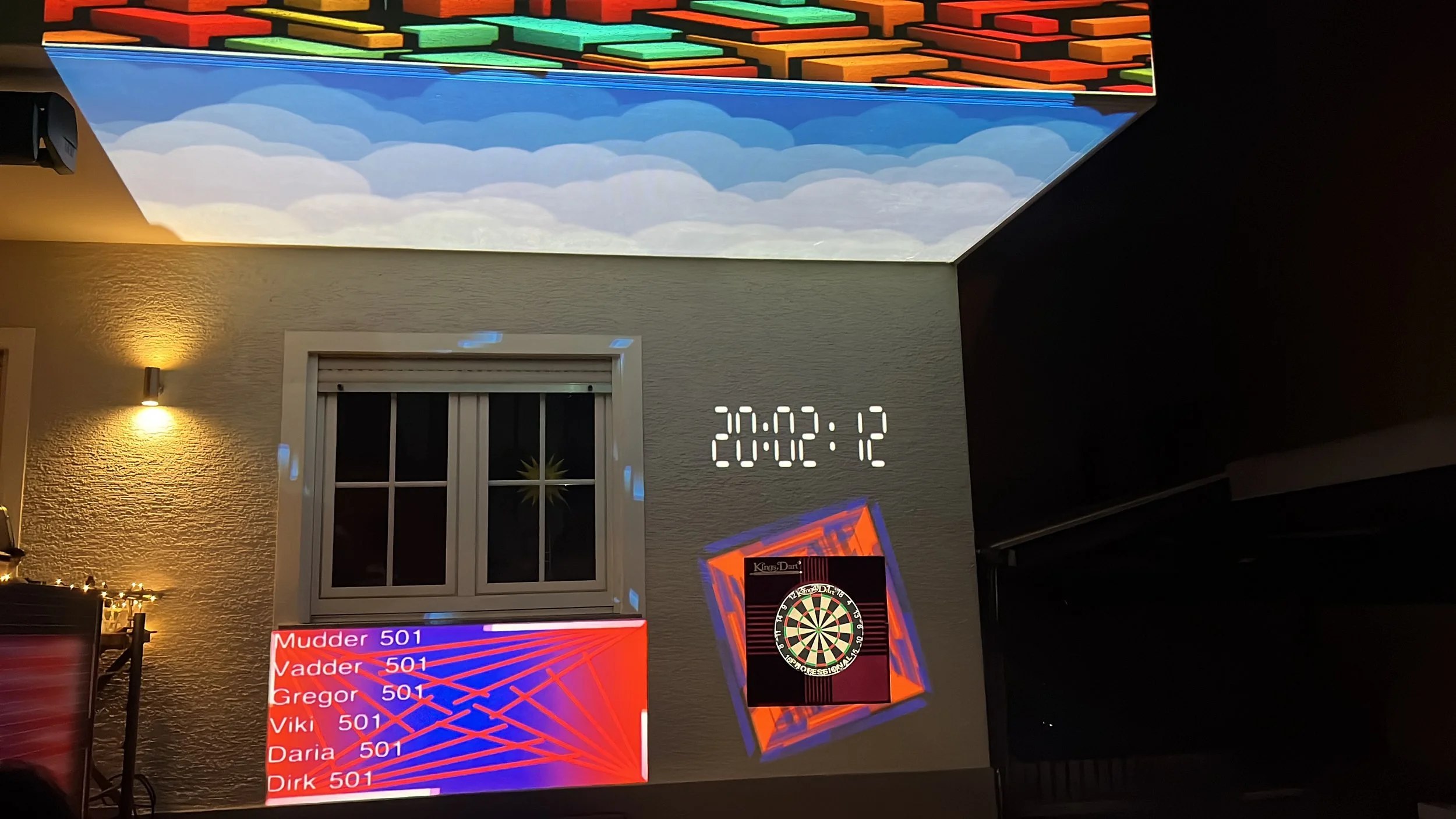 Auf der Außenwand eines Hauses ist eine digitale Uhr, die 20:02:12 anzeigt, zu sehen. Im Hintergrund blickt man durch ein Fenster mit geschlossenen Rollläden. Vor dem Haus hängt an der Wand eine Dartscheibe. Unter der Dartscheibe sind zwei Bildschirme, auf denen Spieler mit den Namen Mudder, Vadder, Gregor, Viki, Daria und Dirk mit der Zahl 501 in blau und rot aufgelistet sind. Die Decke ist mit einer Farbound-Projektionskarte versehen, die Wolkenmuster zeigt.