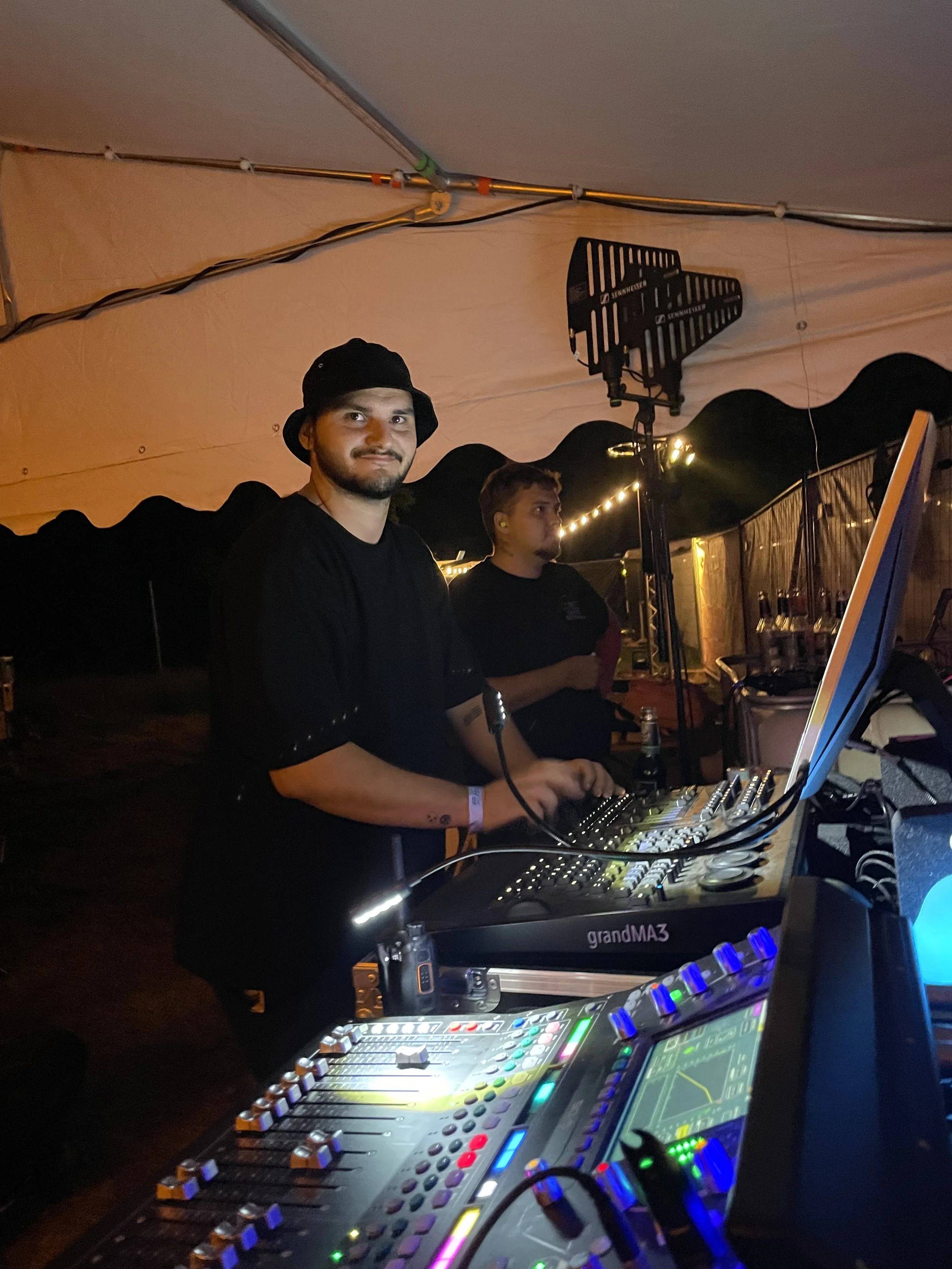 Ein DJ mit schwarzen T-Shirt und Hut steht hinter einem DJ-Controller bei einer Party im Zelt, im Hintergrund sind Lichter und eine weitere Person.