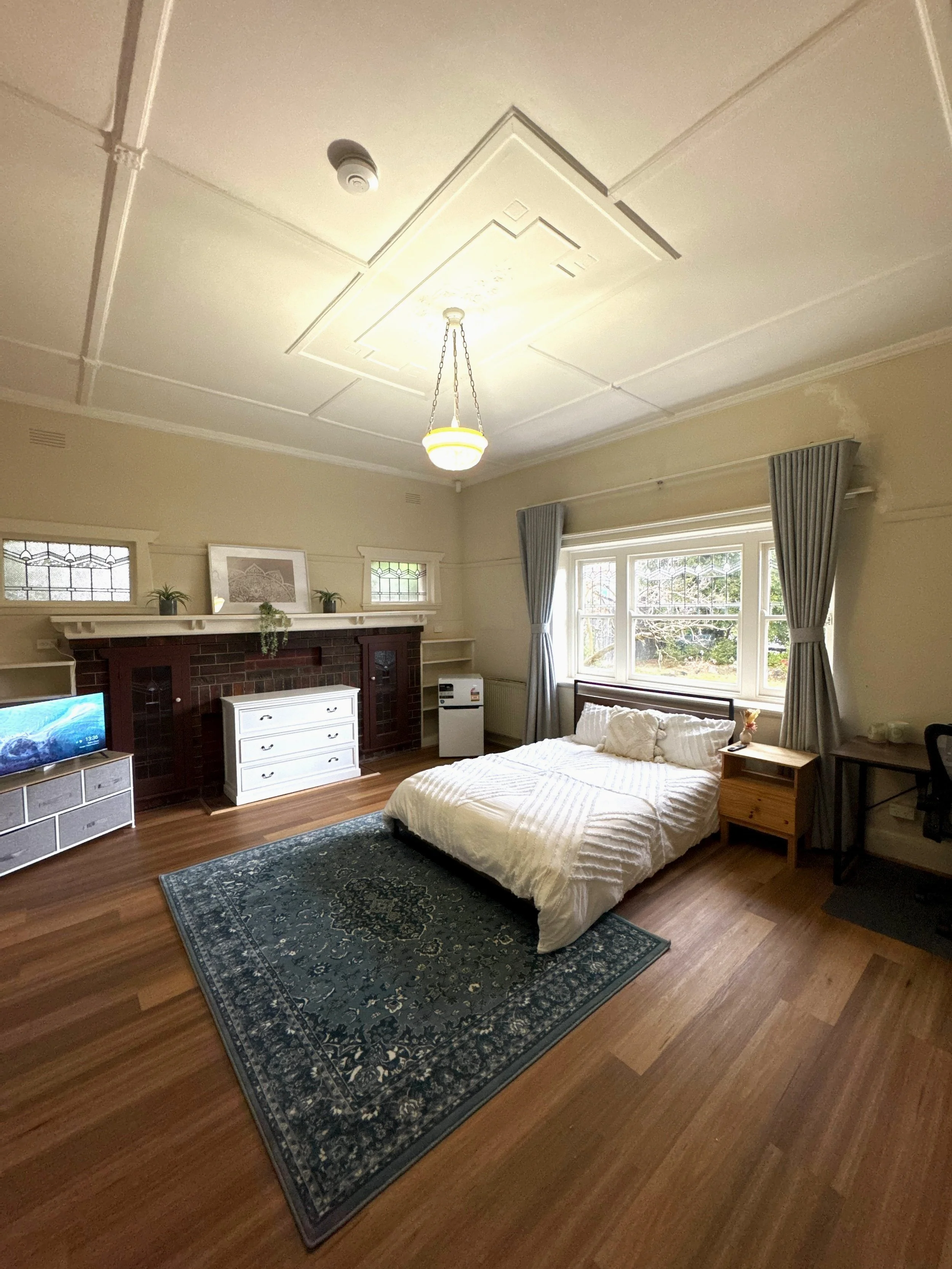 A converted rooming house with a bed, nightstand, dresser, small TV, and window with curtains. The room has a wooden floor and a decorative area rug.