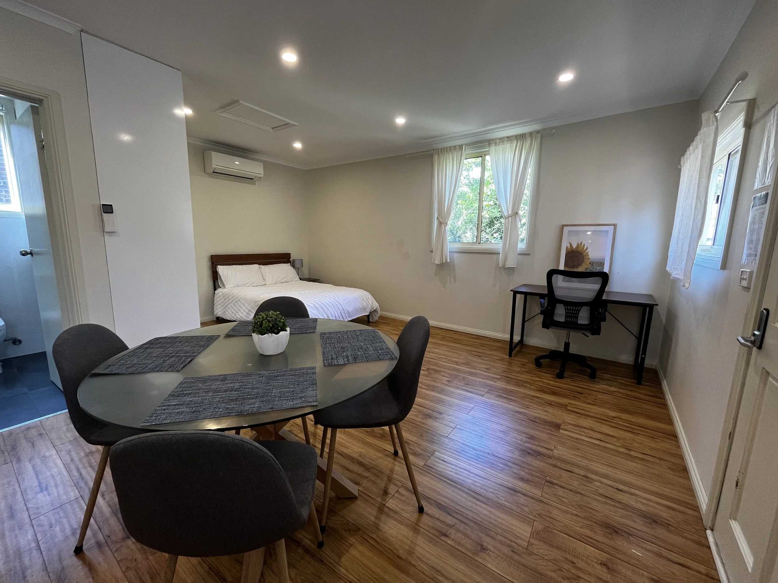 Converted rooming house room a wooden floor, dining room, a bed, and a small workspace near the windows.