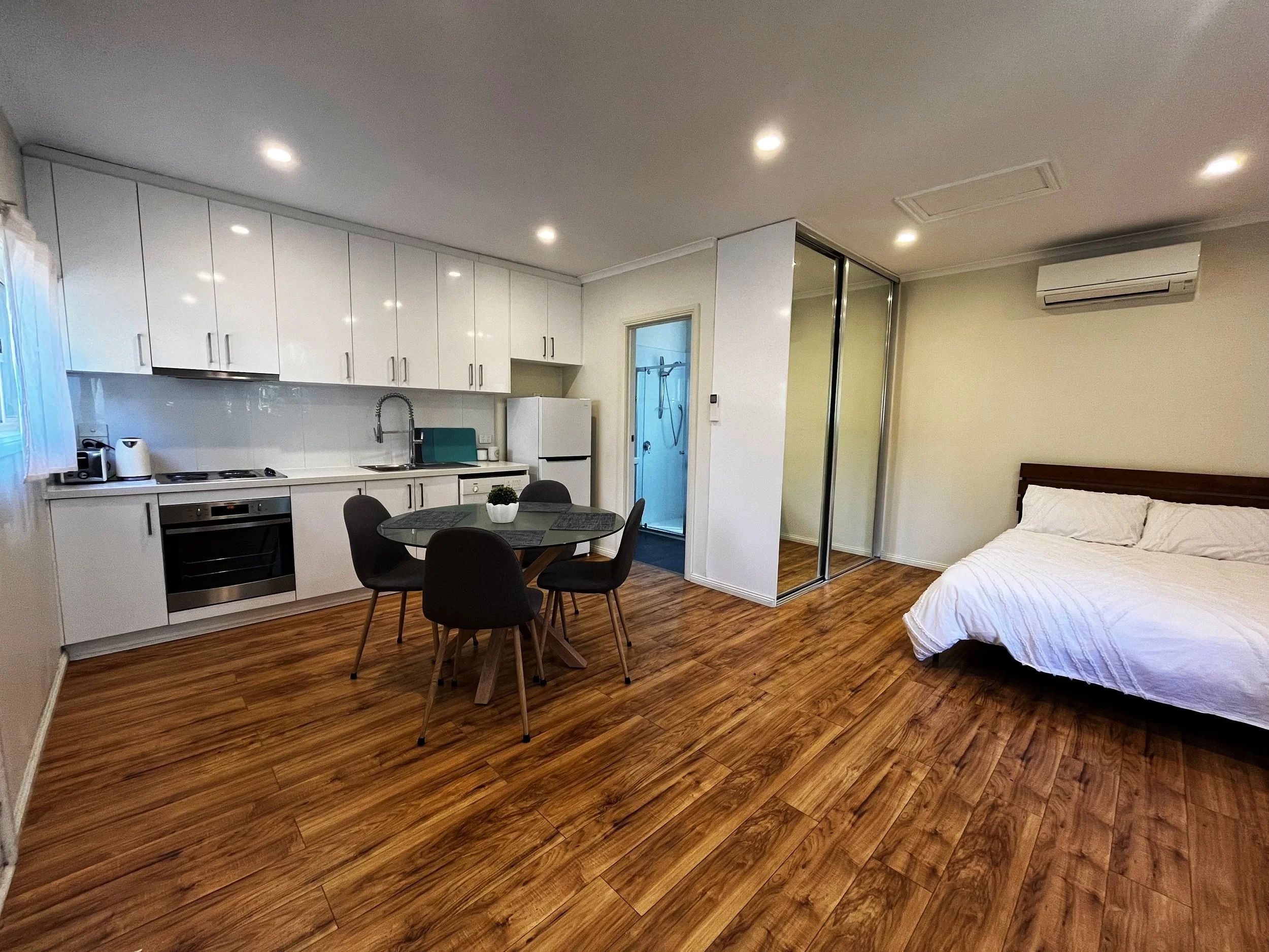 Converted rooming house with white kitchen, dining room, bed with white bedding, hardwood floors, and mirrored closet doors.