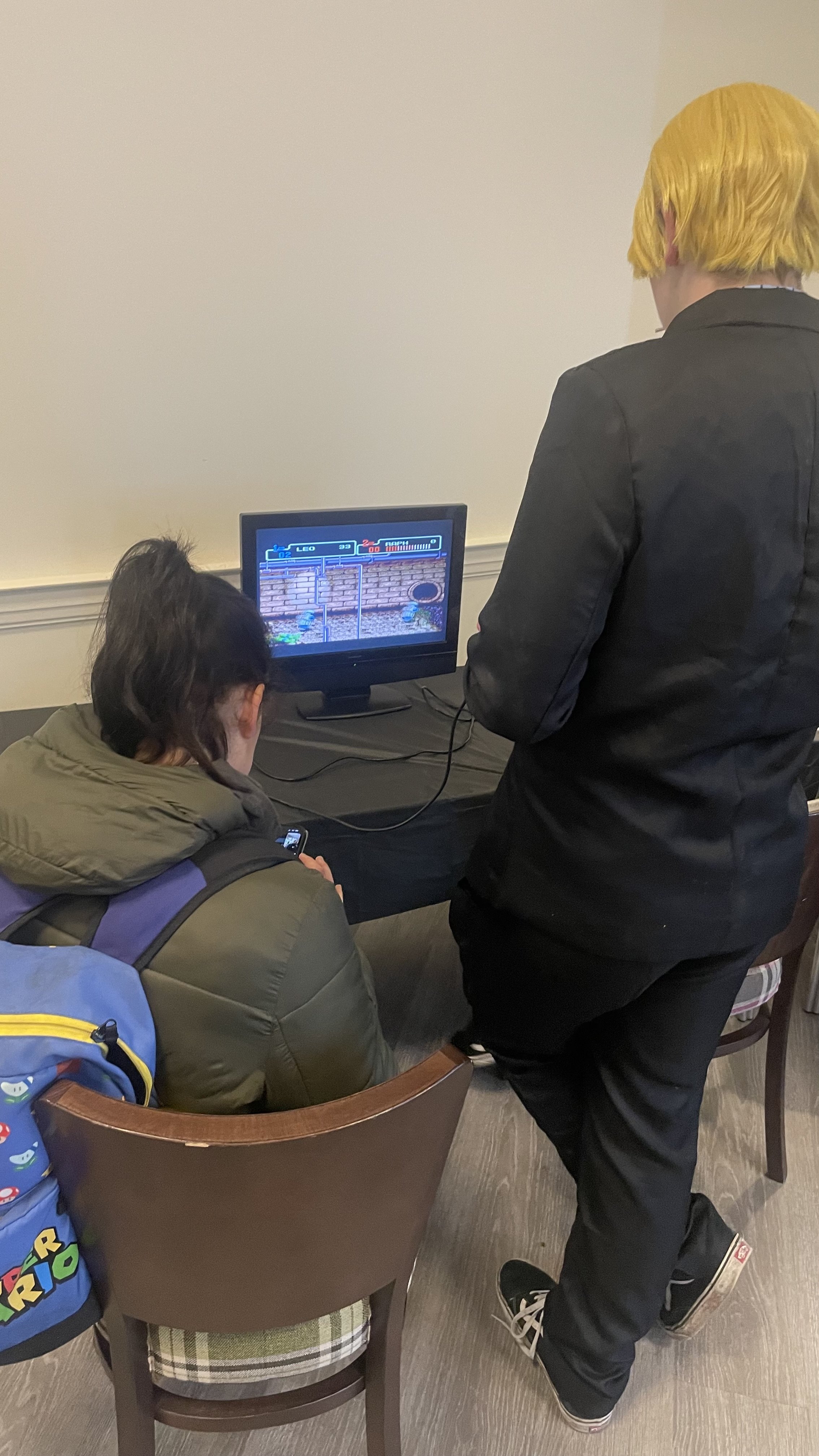 A person with yellow hair stands next to a seated person playing a video game on a monitor, with a backpack on the chair.