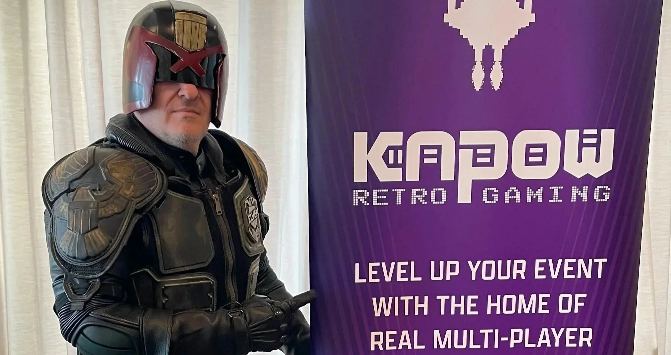 A person dressed in a futuristic, armored costume with a helmet, standing next to a purple banner that reads 'KAPOW RETRO GAMING' and 'LEVEL UP YOUR EVENT WITH THE HOME OF REAL MULTI-PLAYER'.