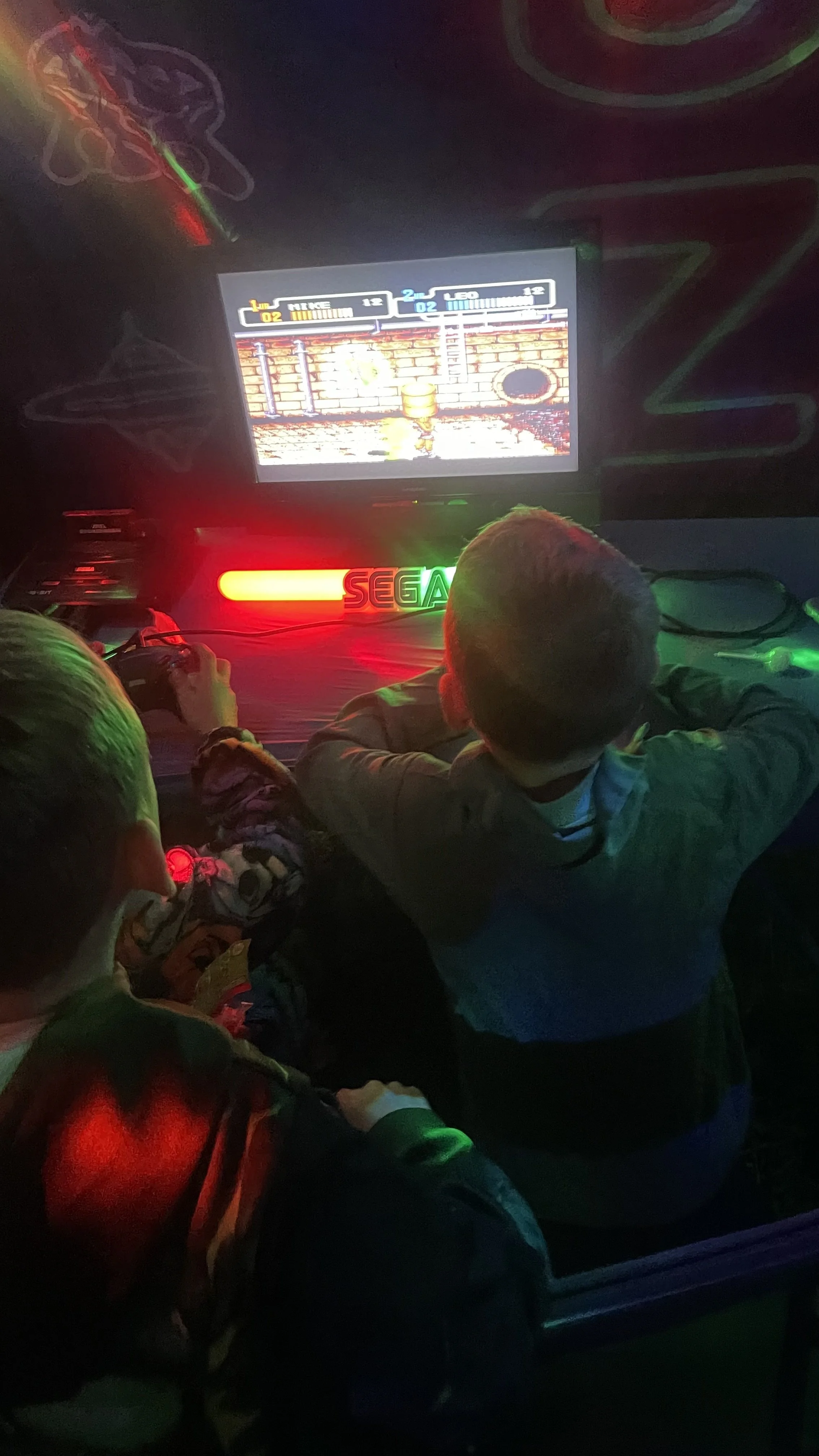 Two children playing an arcade game at a Sega arcade machine with a neon sign, in a dark room with colorful lighting and animated wall decorations.