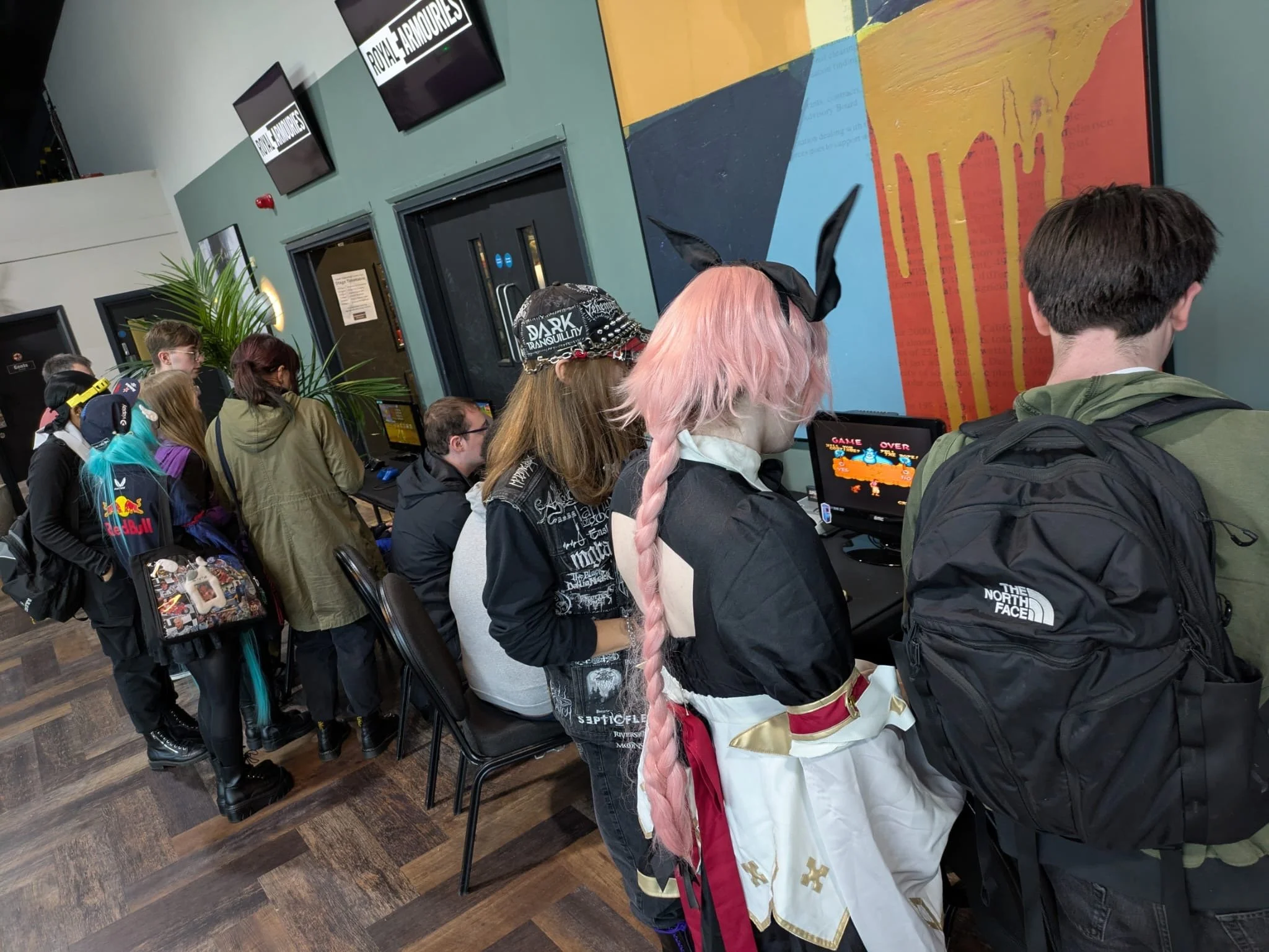 Line of people waiting to play arcade games at an indoor gaming venue, with some dressed in costumes and casual wear, with a colorful mural on the wall and large monitor screens displaying the venue's name.