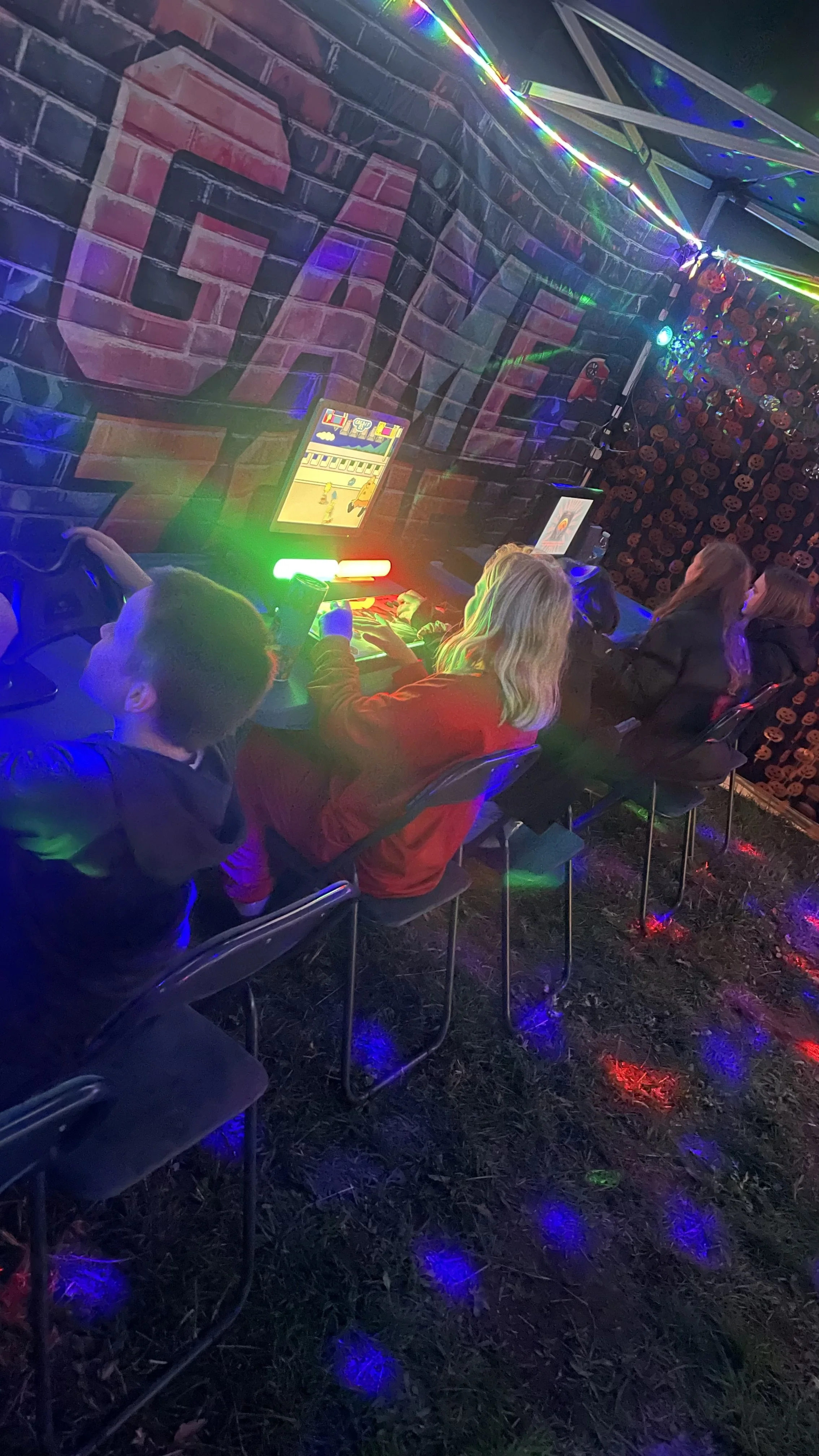 People sitting on chairs playing arcade games at a decorated event with colorful lights and Halloween-themed decorations.
