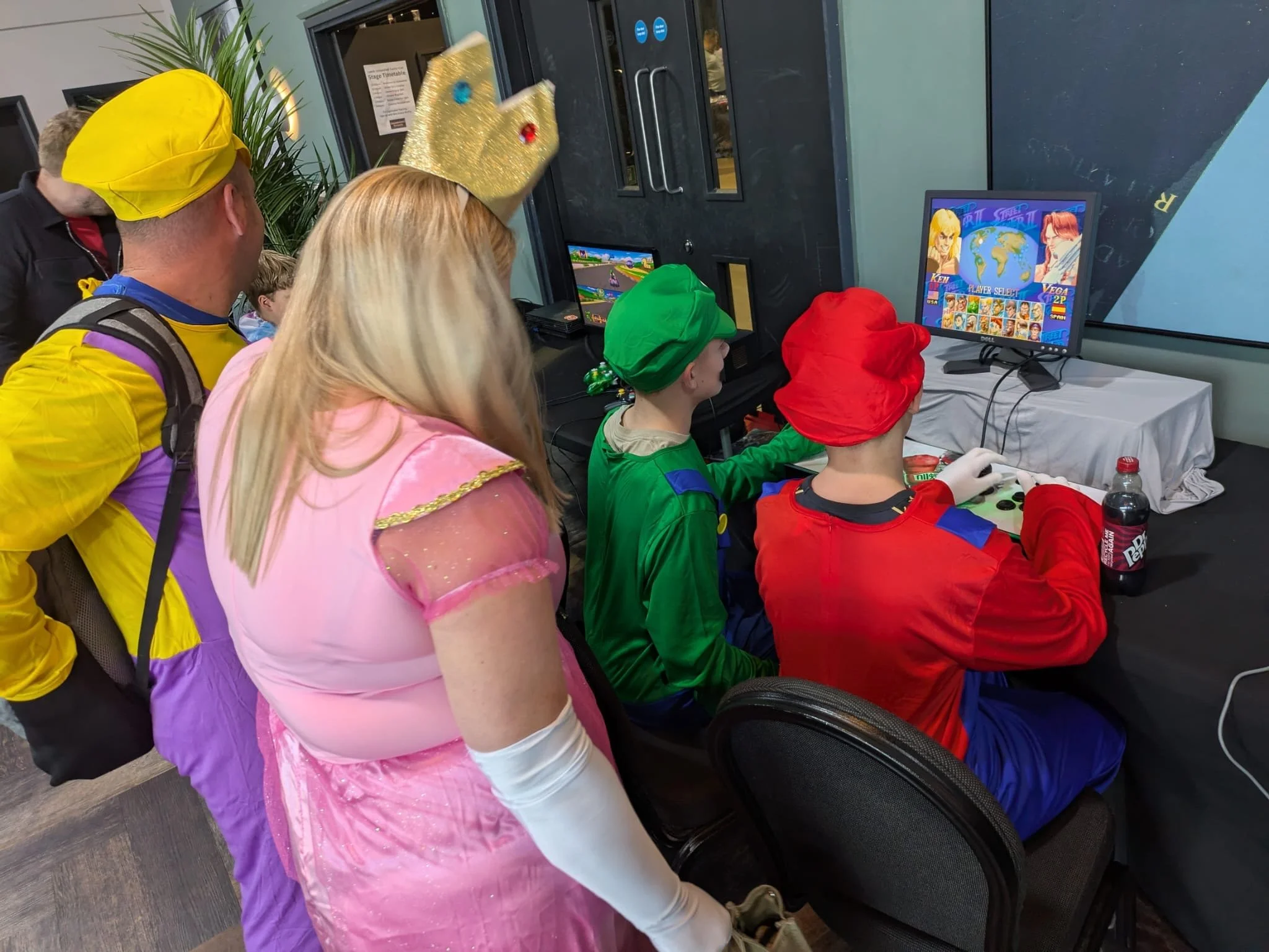 Group of people dressed in colorful costumes gathered around two video game consoles, playing a fighting game at an indoor event.