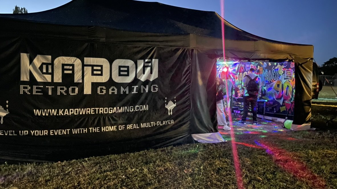Outdoor event tent with a sign that reads 'KAPOW RETRO GAMING' and a website URL. Inside, people are playing arcade games with colorful graffiti-style art on the walls. The area is decorated with pink laser lights and balloons, and it's evening.