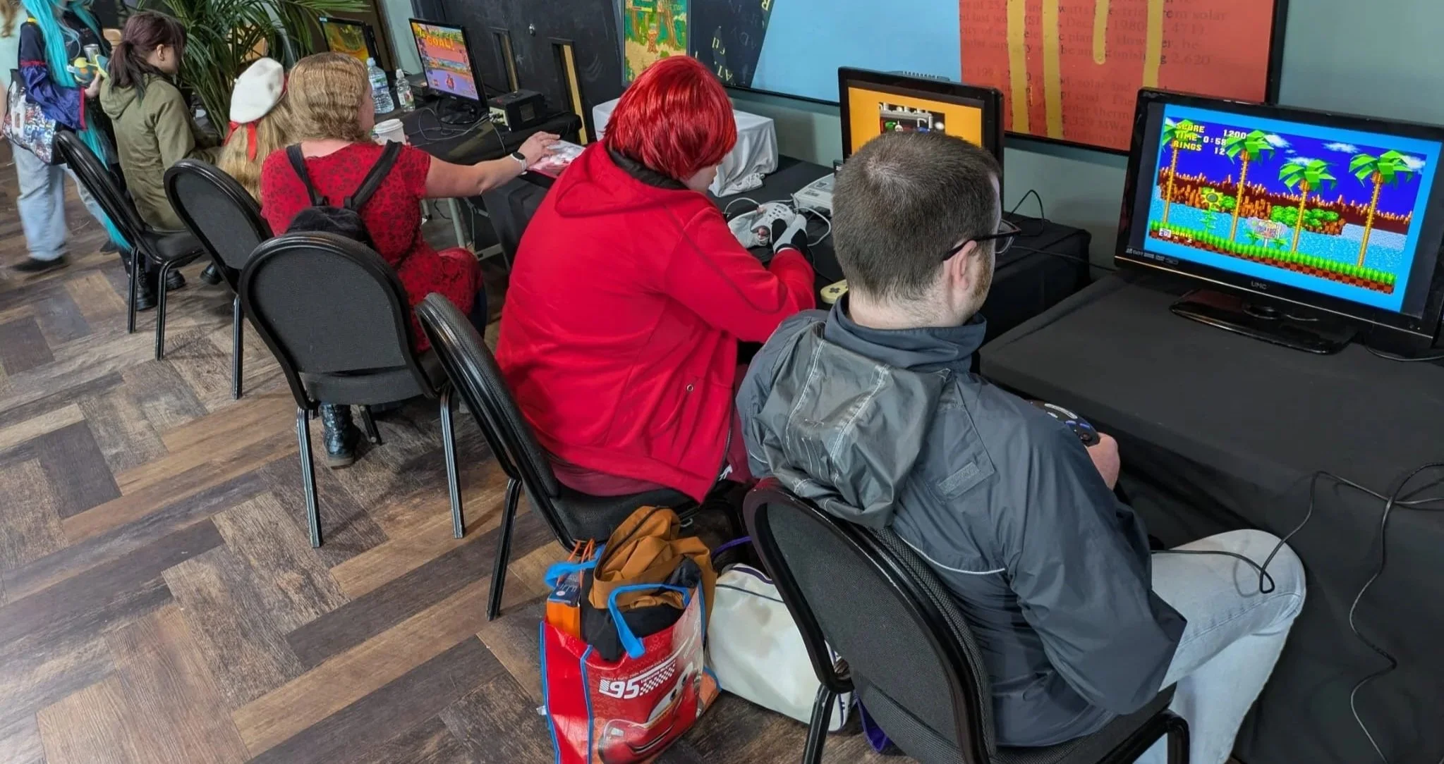 People playing video games on arcade machines at a gaming event, with several individuals seated and focused on screens displaying Sonic the Hedgehog.