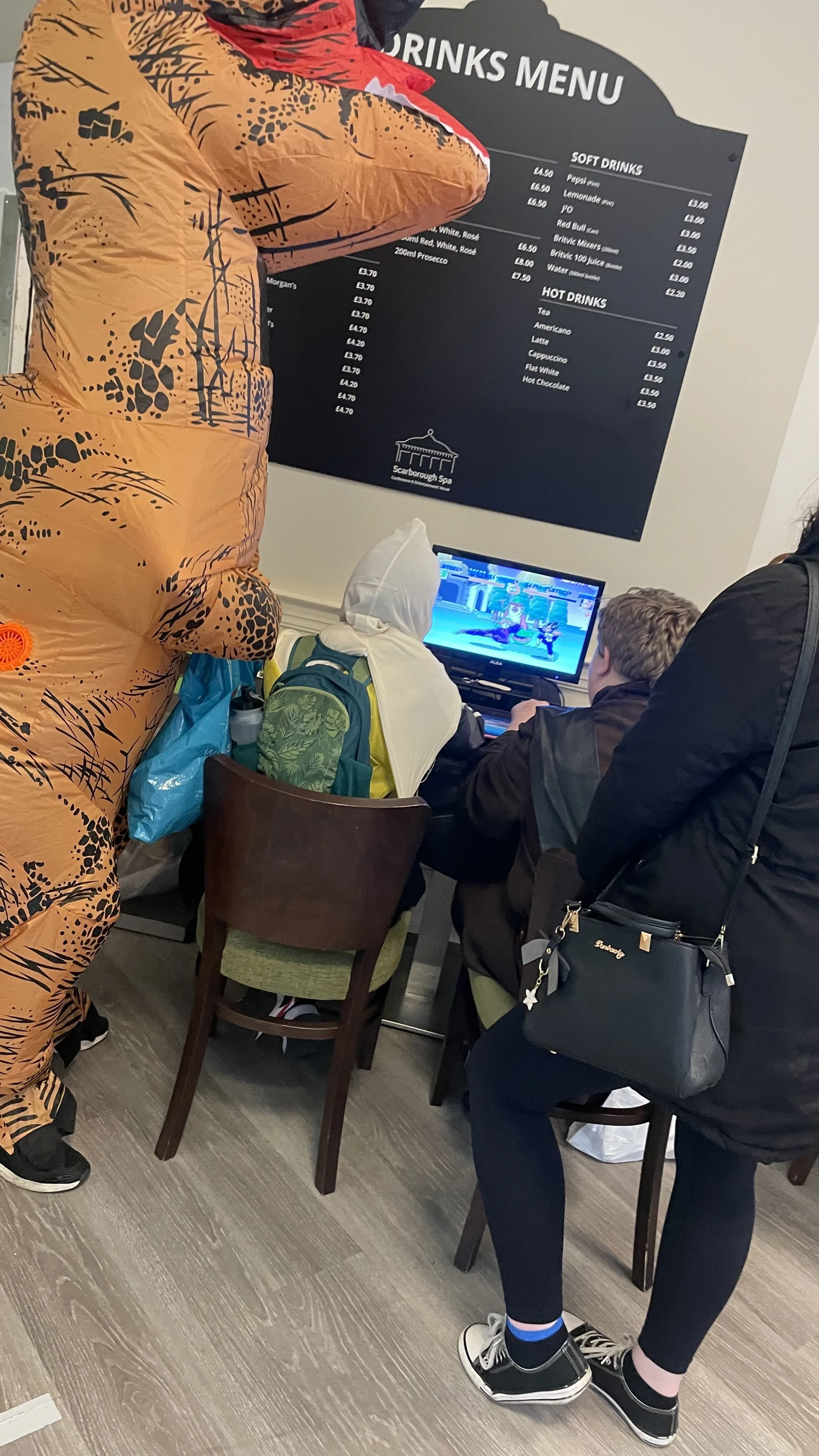 Group of people in a room, one wearing an animal costume, all focused on a computer screen showing a fighting game, with a drinks menu on the wall in the background.