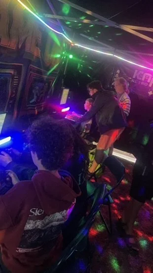 People playing arcade video games in a dimly lit arcade with colorful laser lights and neon signs.