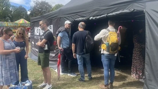 Group of people gathered outside a black tent at an outdoor event or fair, some looking at their phones, with colorful tents and trees in the background.