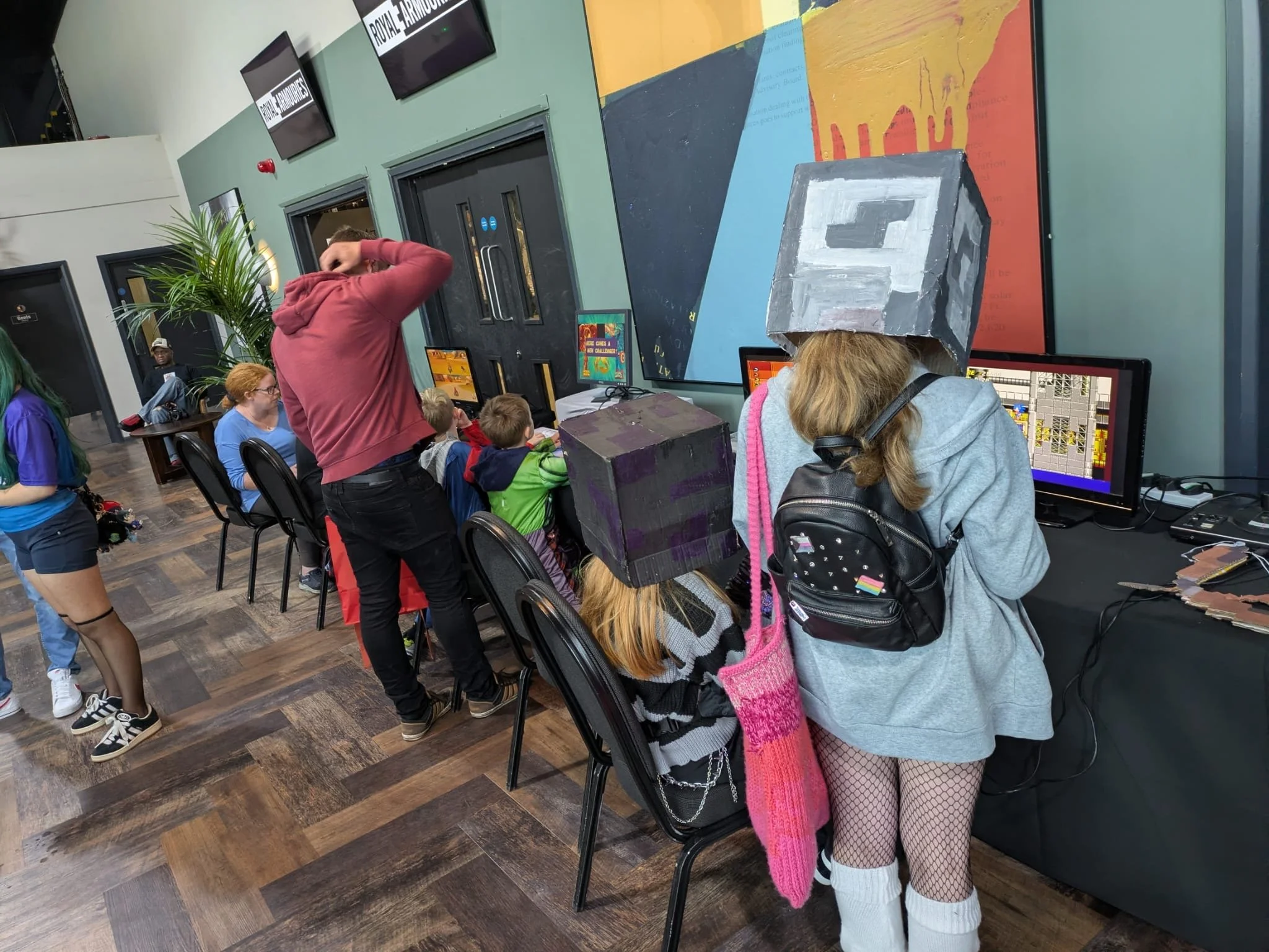 People at a gaming event, including individuals dressed in Minecraft-themed costumes with pixelated boxes on their heads, playing video games on computers, and others watching. The room has wooden flooring, colorful walls, large monitors, and gaming posters.