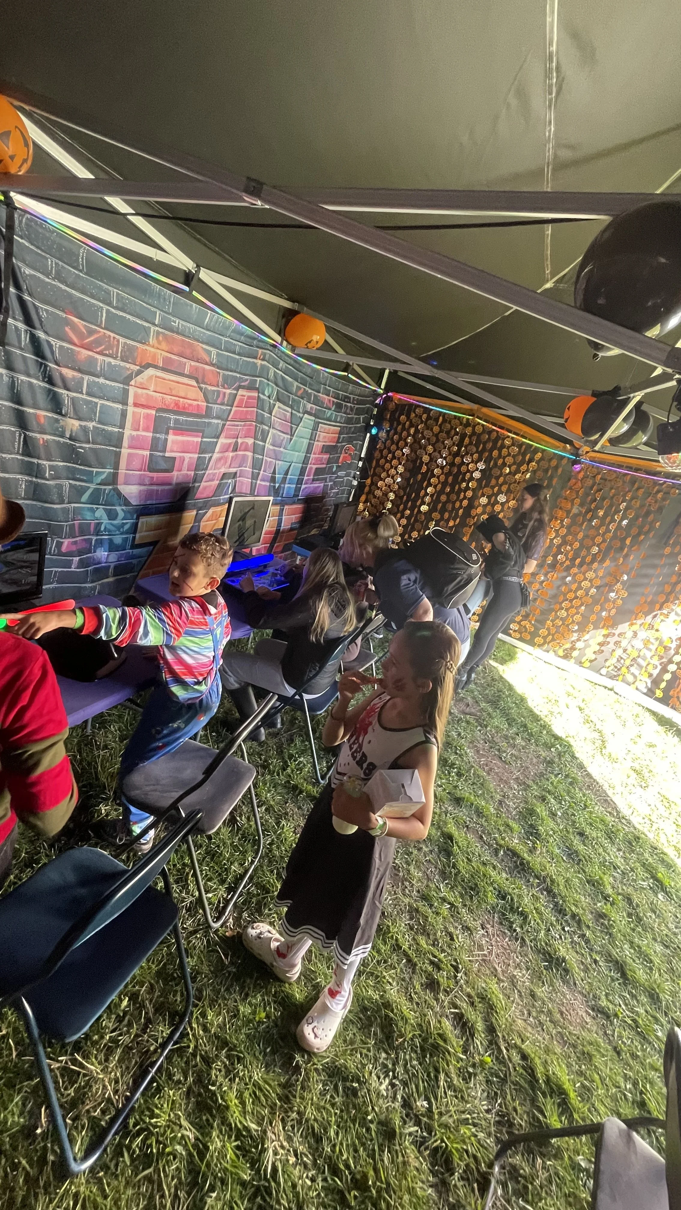 Outdoor game arcade at night with children playing and a girl eating in the foreground, decorated for Halloween with pumpkins and strings of lights, and with a colorful graffiti-style wall in the background.