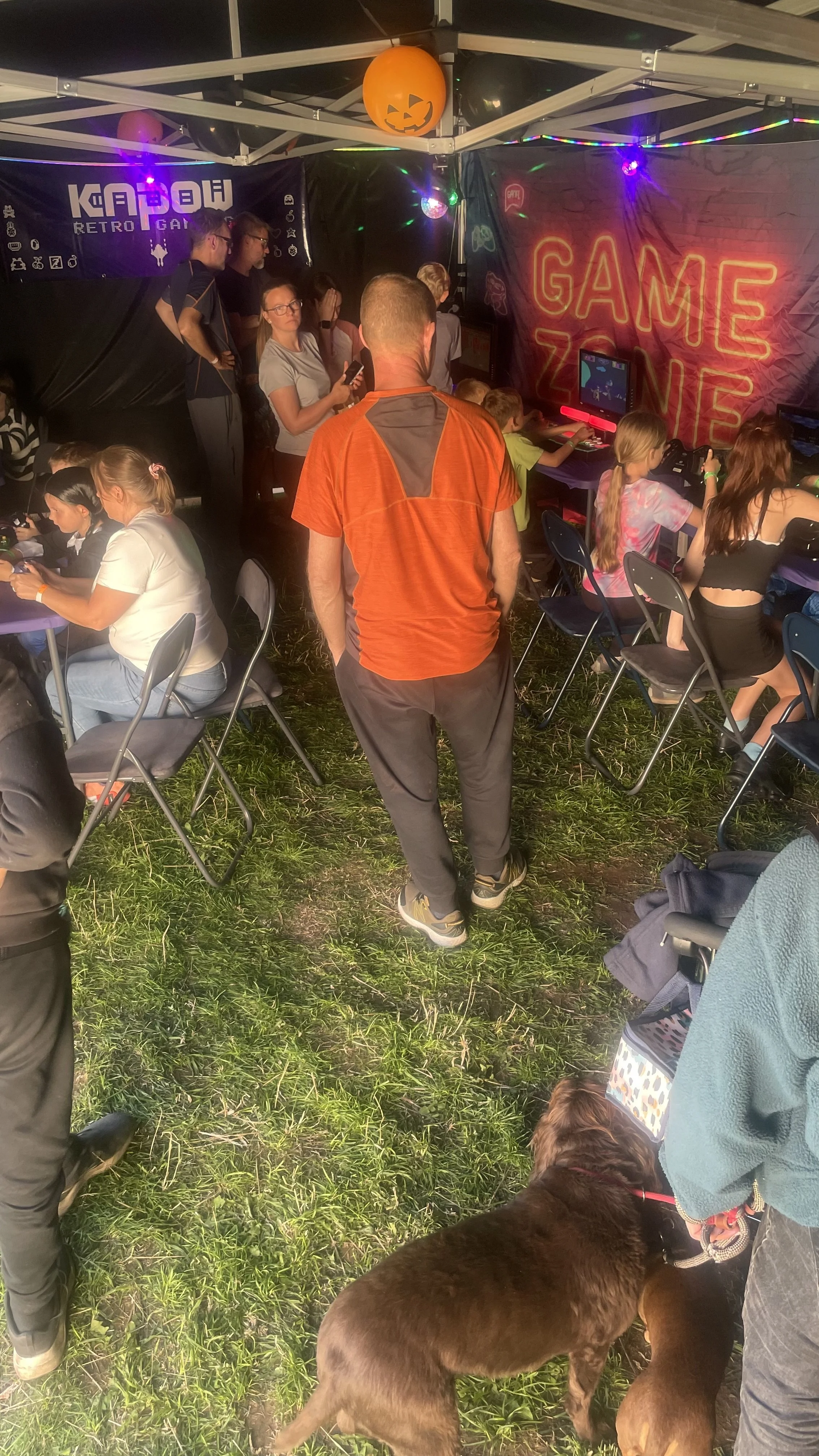 People at a gaming event under a tent decorated with Halloween-themed decorations, including a pumpkin-shaped hanging lantern. Several children are playing video games on screens, with a neon sign that says 'GAME ZONE'. Adults are watching or waiting, and a dog is on a leash near the bottom right of the image.