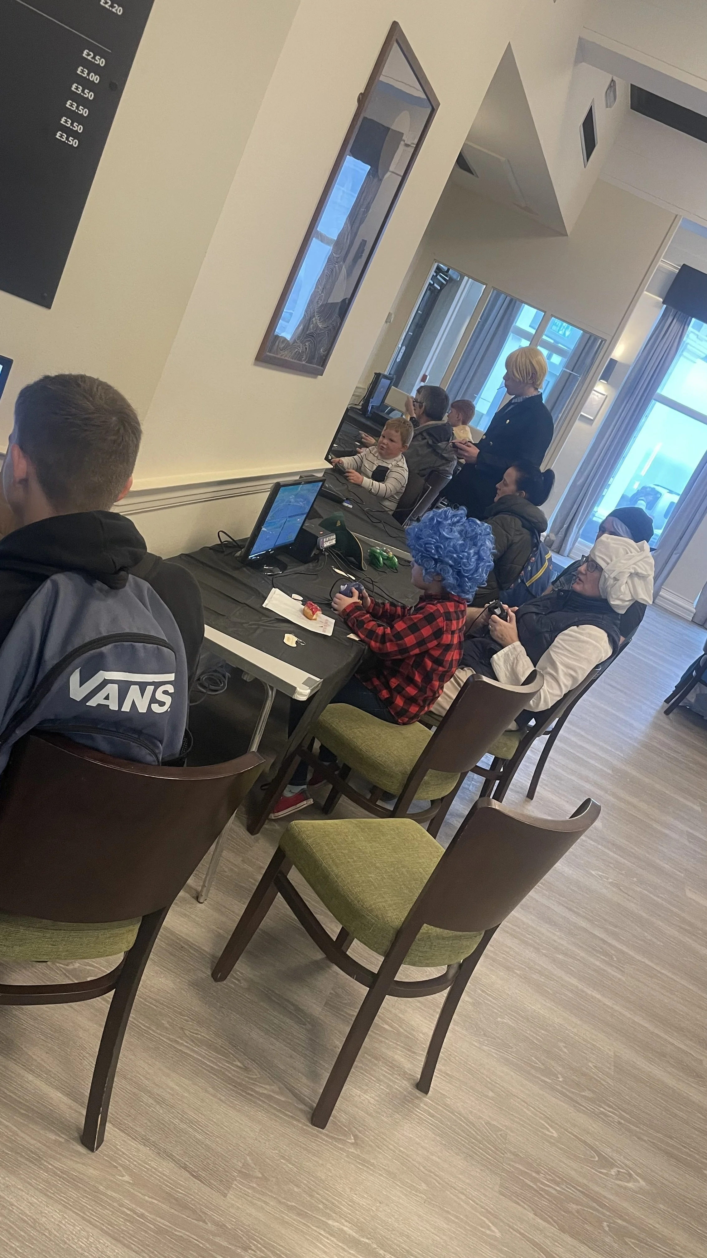 People playing video games in a cafe or gaming center, with a child wearing a blue wig and another dressed as a chef.