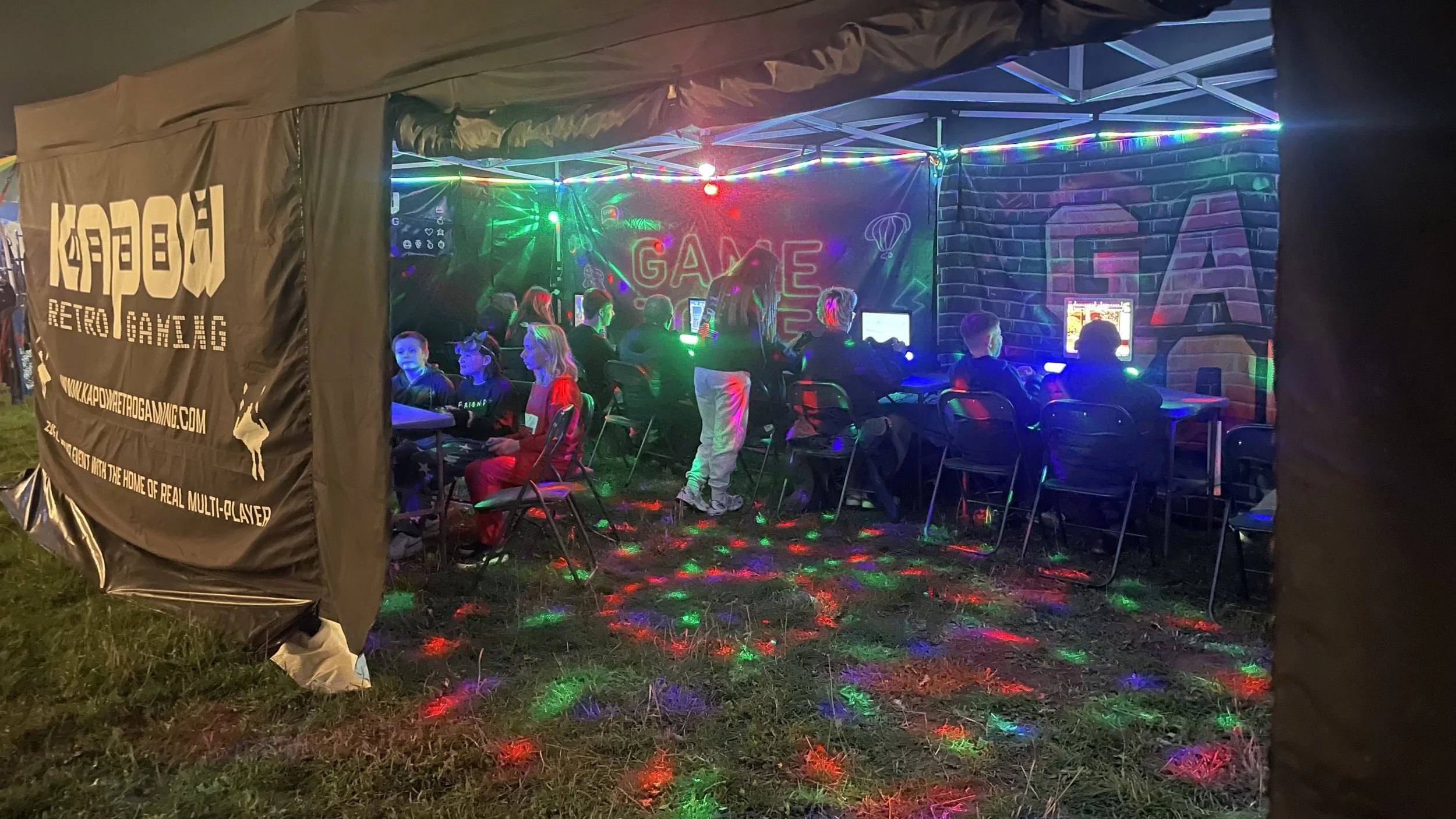 Kids playing video games inside a tent with colorful neon lights and decorations.