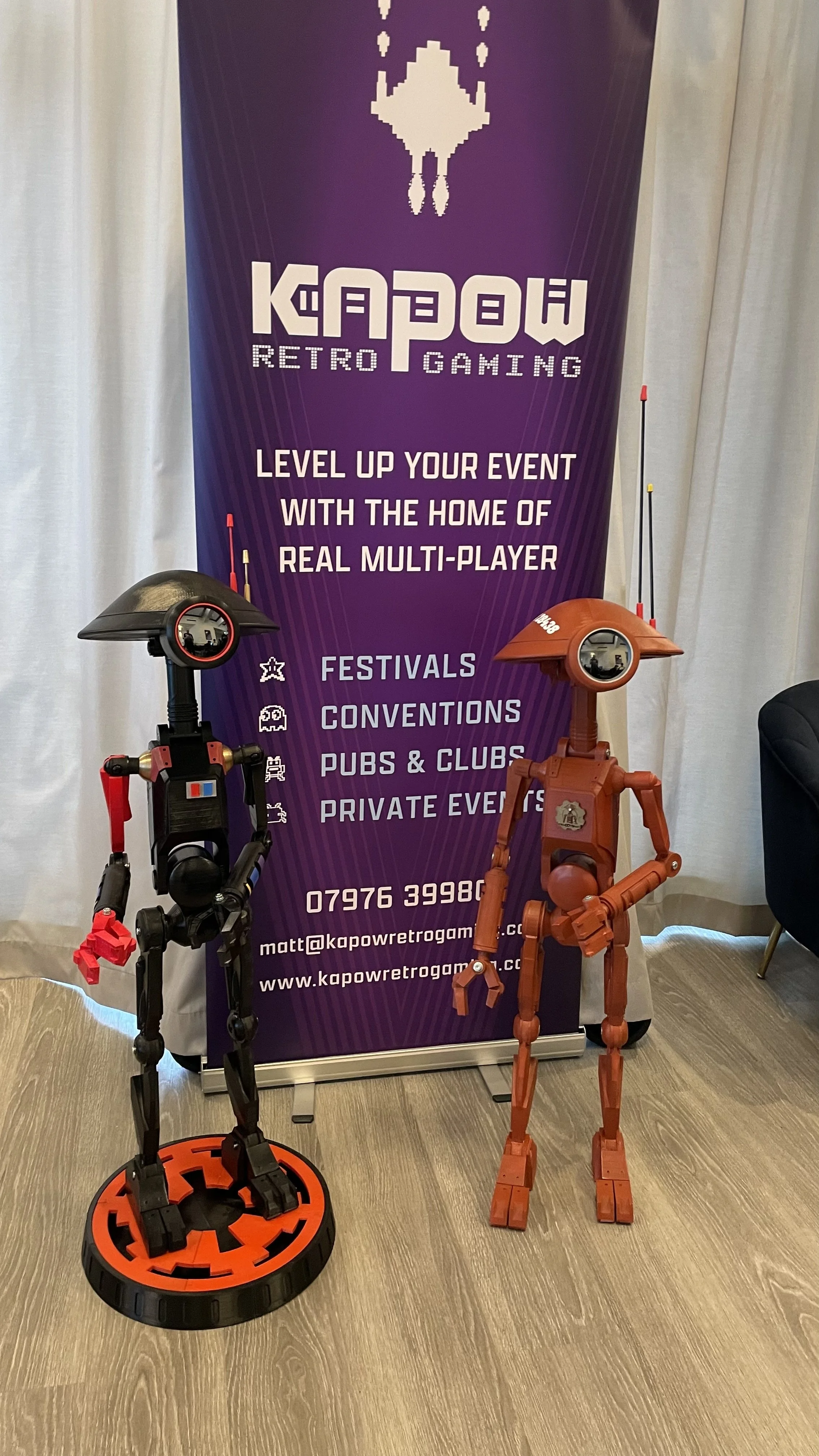 Two robot figures, one black and red and one brown, in front of a purple Kapow Retro Gaming banner with text promoting multi-player gaming at festivals, conventions, pubs, clubs, and private events.