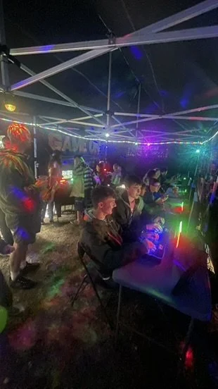 Children playing video games at a night event under a canopy with colorful lights.