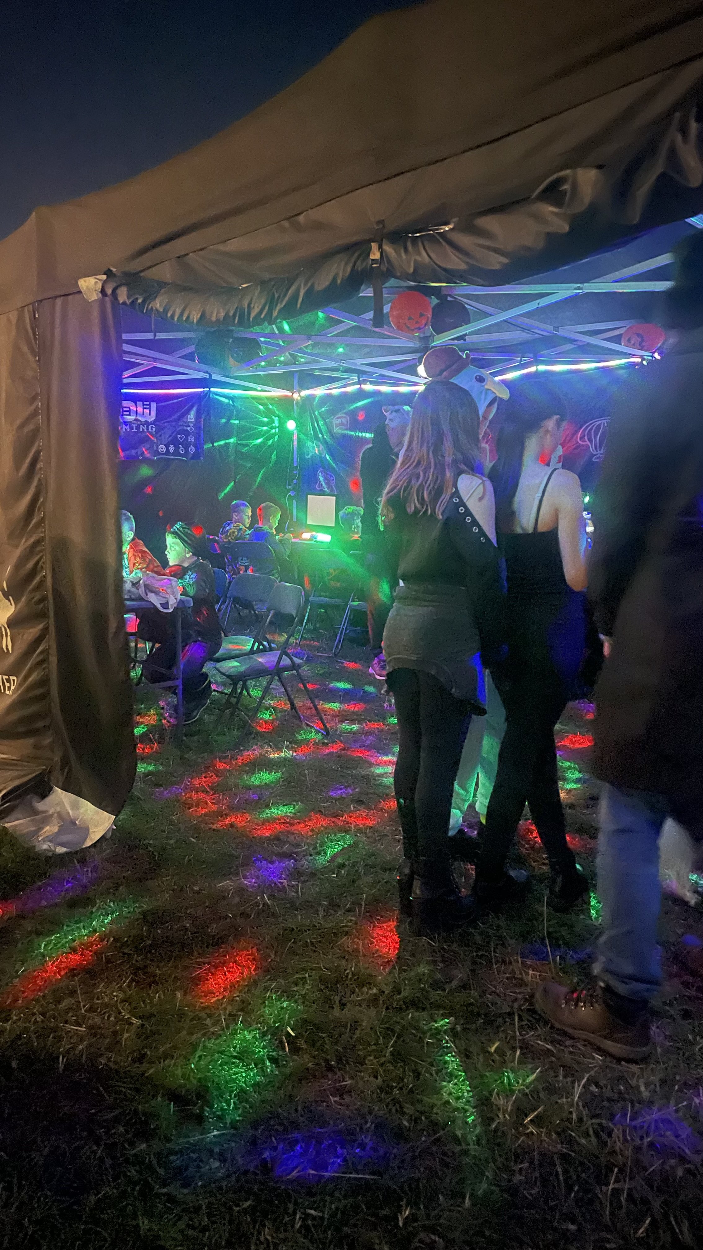 A group of people at a Halloween-themed indoor event, with colorful lights and decorations, some wearing costumes including a snowman hat, some sitting at a table, and others standing and walking around.