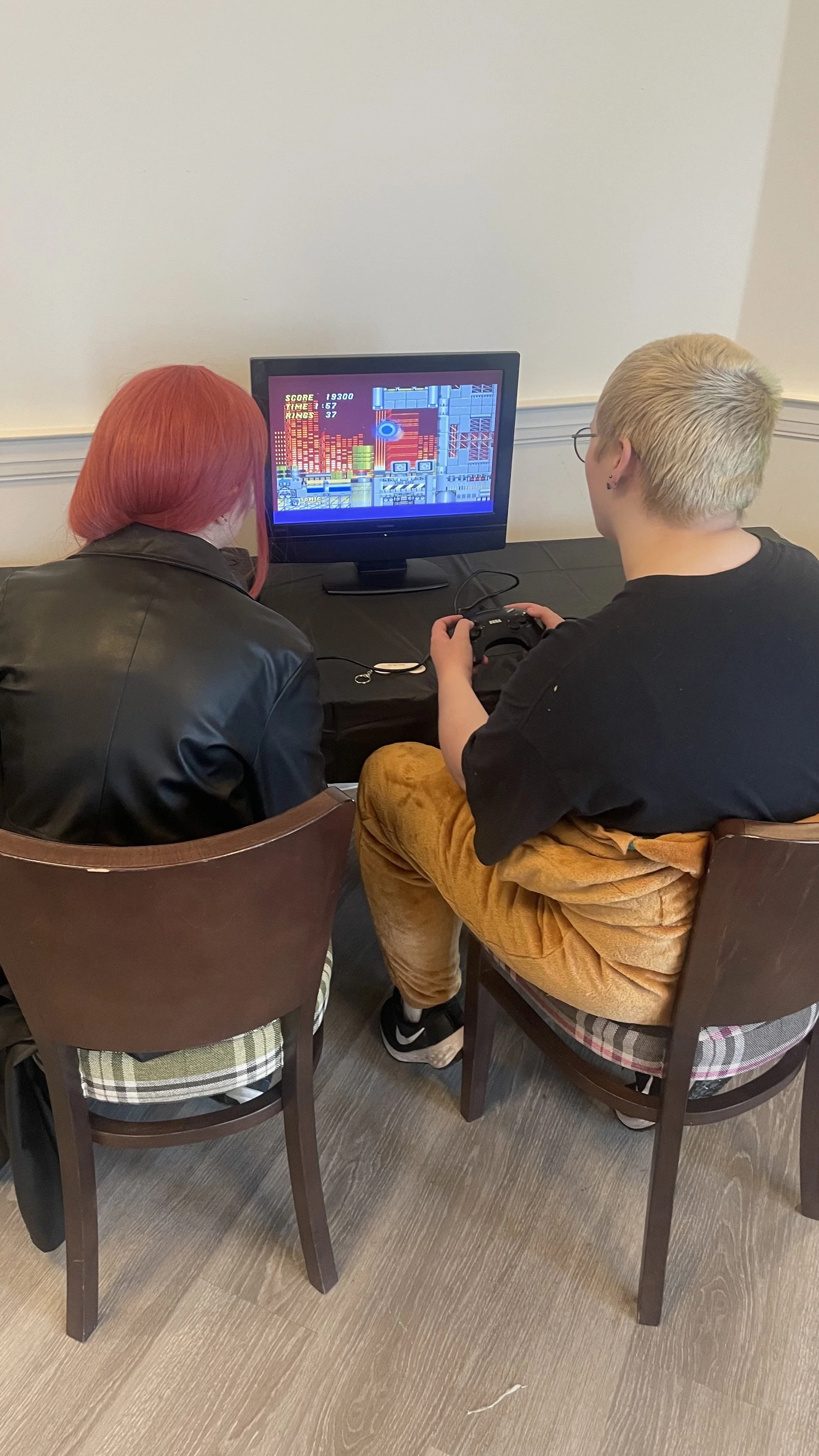 Two people sitting in chairs and playing a video game on a small TV, with one person holding a game controller and the other person watching.