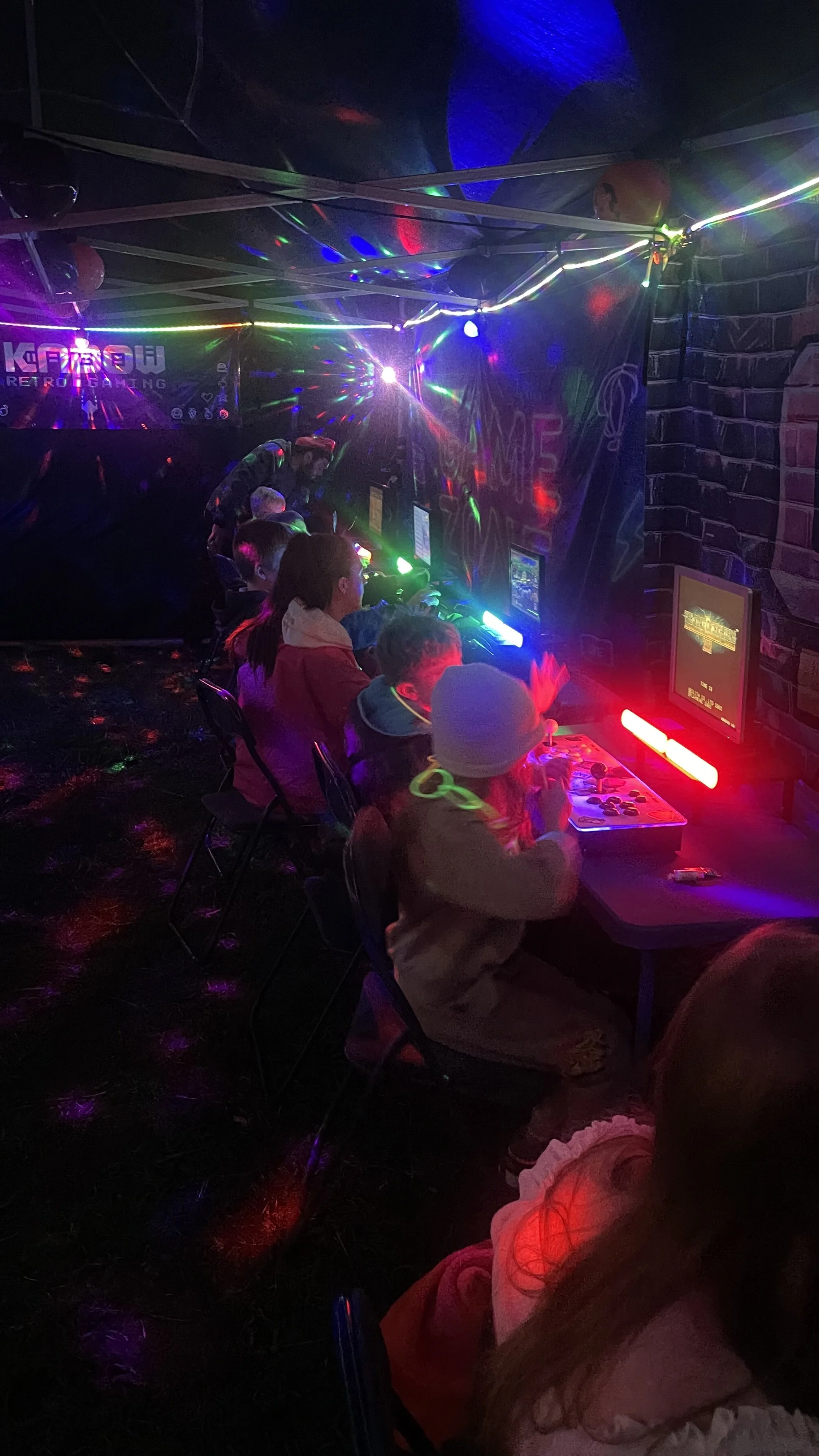 Children playing arcade games in a dimly lit room with neon lights and disco reflections.