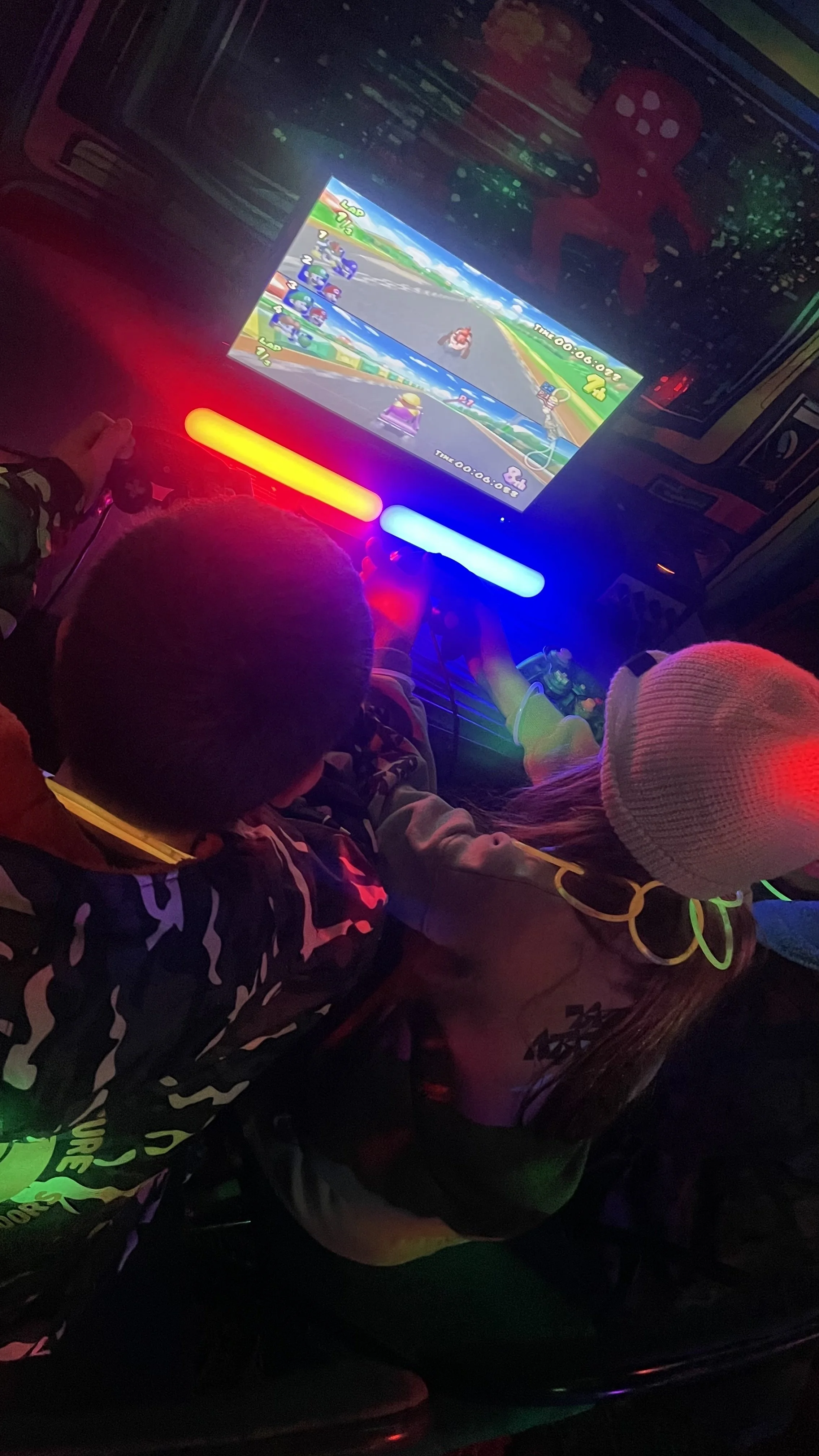 Two children playing a racing video game in a dark room with colorful LED lights. A screen displays a Mario Kart game with characters racing on a track.
